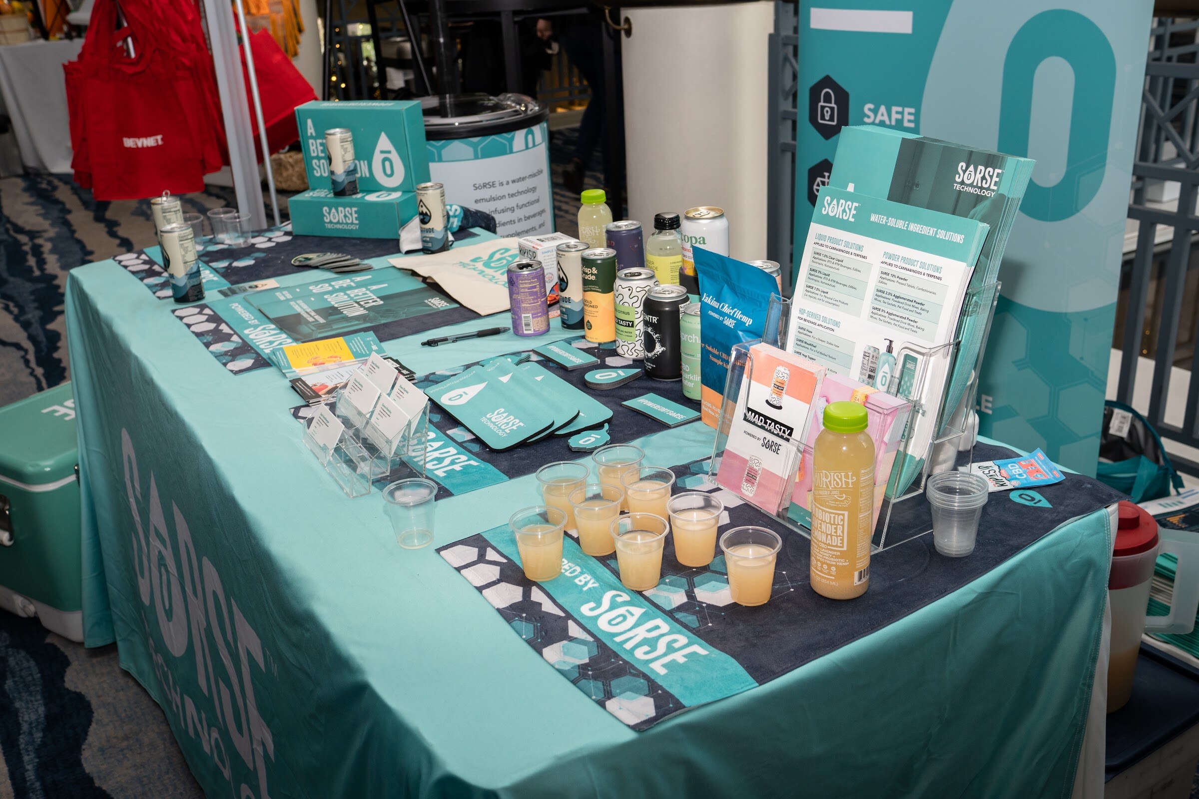A teal tablecloth printed with Sorse holds brochures, small sample cups of orange juice, cans and a bottle in front of a tall Sorse banner.