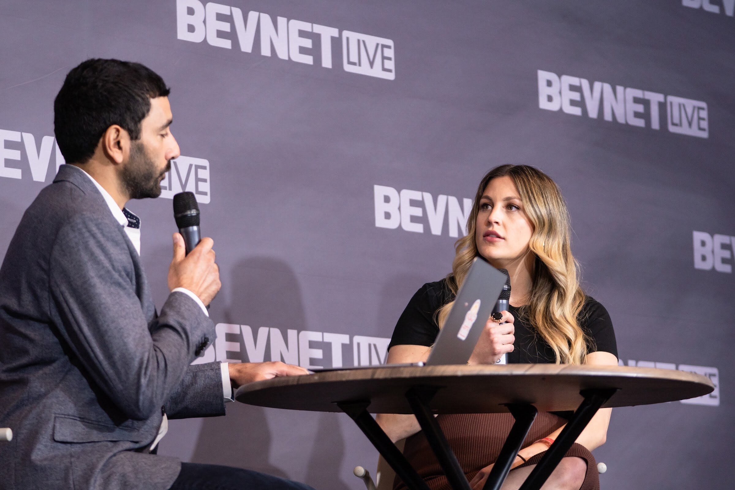 A man in a gray blazer holding a microphone sits across a round table from a woman with long wavy hair and a laptop in front of a BevNET Live backdrop.
