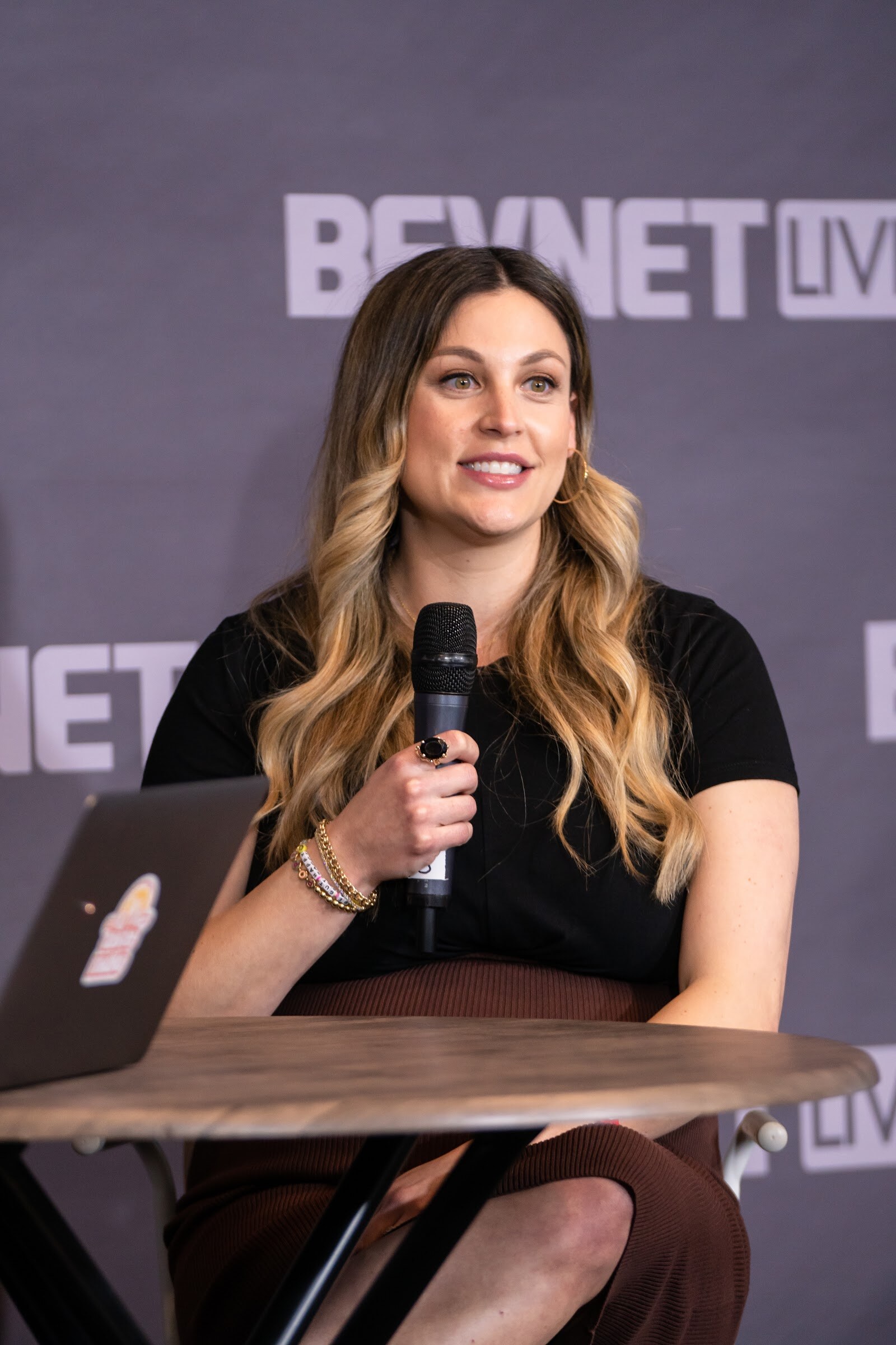 A smiling woman with long wavy hair in a black top holds a microphone at a round wooden table in front of a purple BevNET Live backdrop.