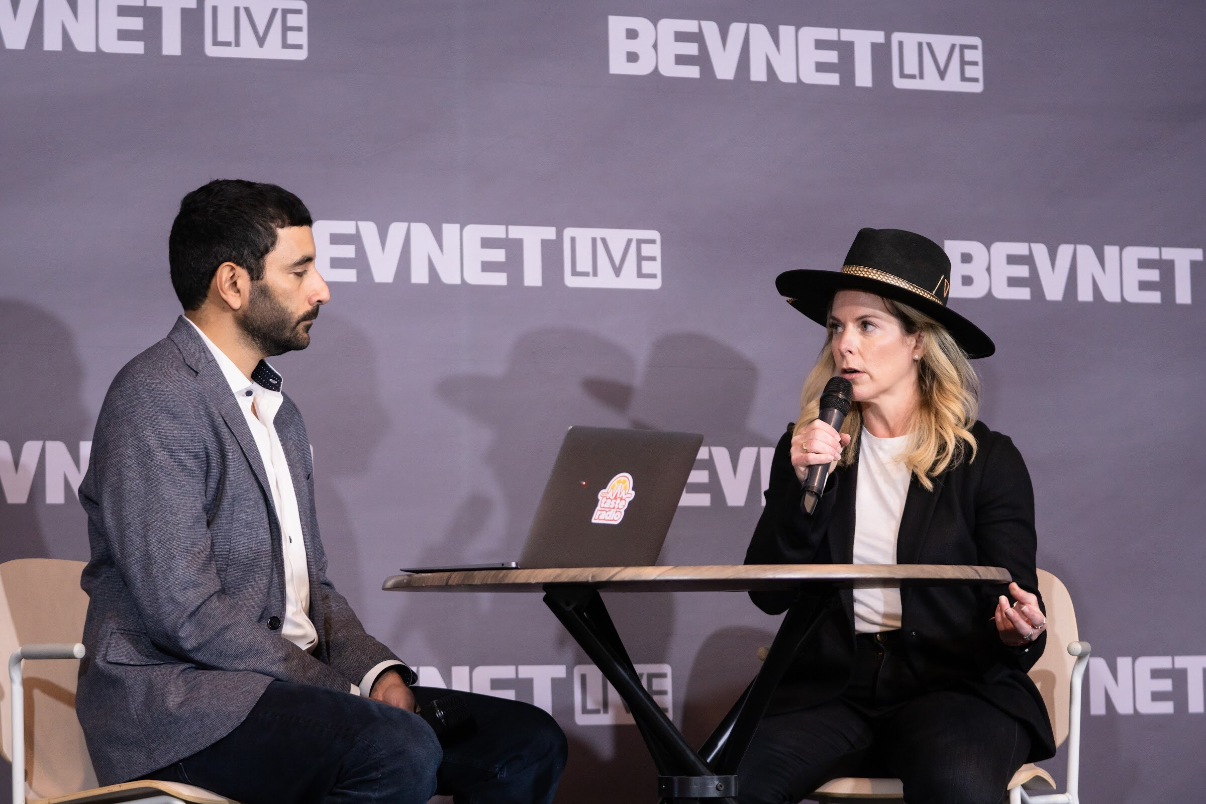 A man in a gray blazer and a blonde woman in a black hat and blazer hold microphones on either side of a round table with a laptop.