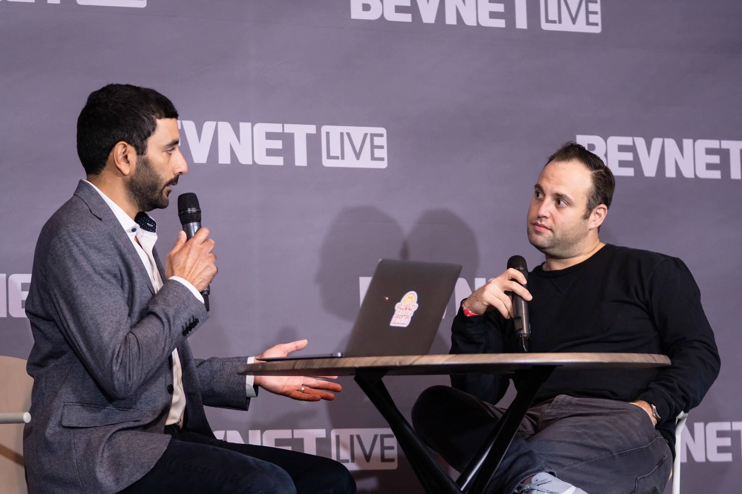 A man in a gray blazer holds a microphone across a round table from a man in a black sweater holding a microphone, in front of a BevNET Live backdrop.
