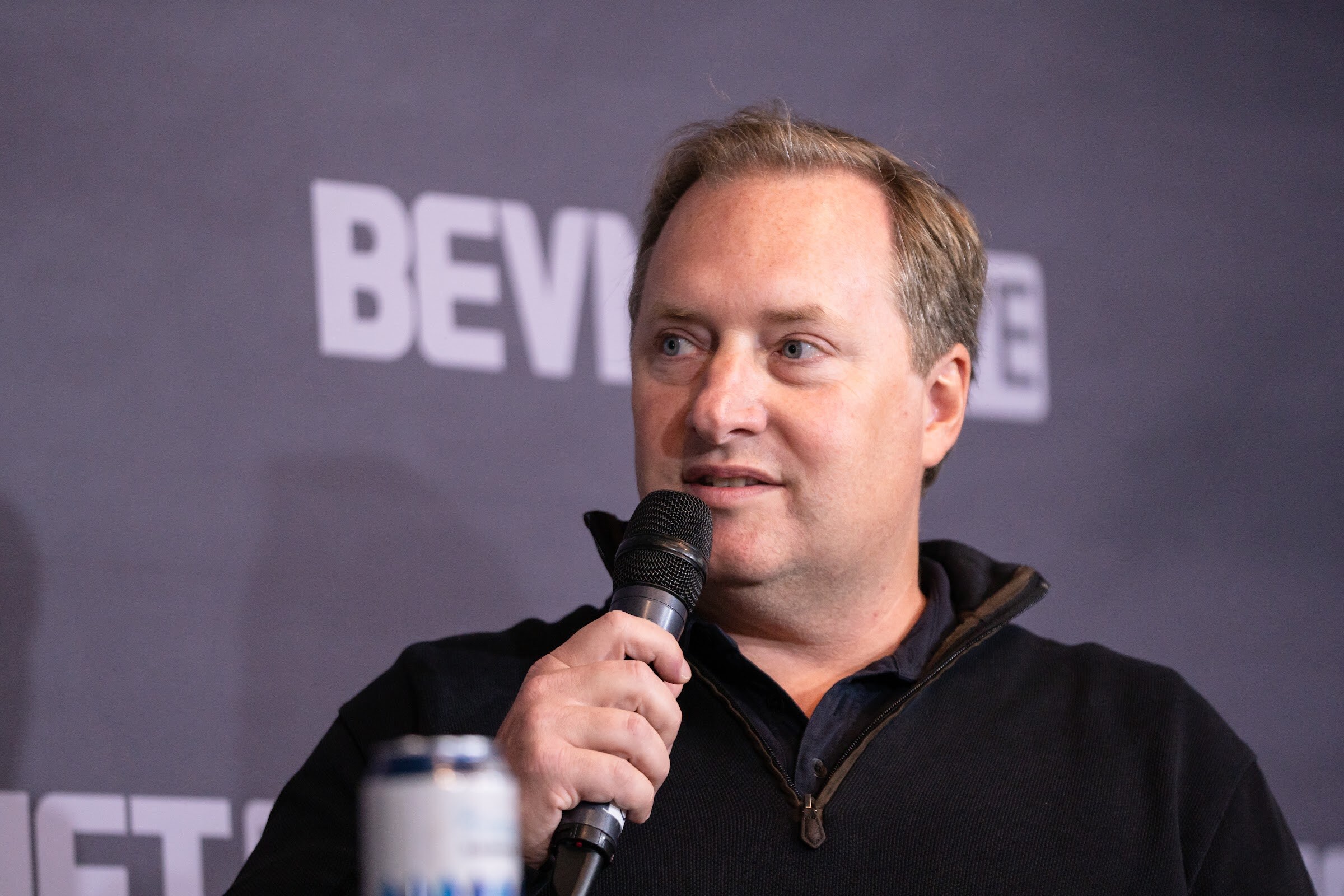 A man in a black quarter-zip sweater speaks into a microphone in front of a purple BevNET Live backdrop, with a can partly visible at the lower edge.