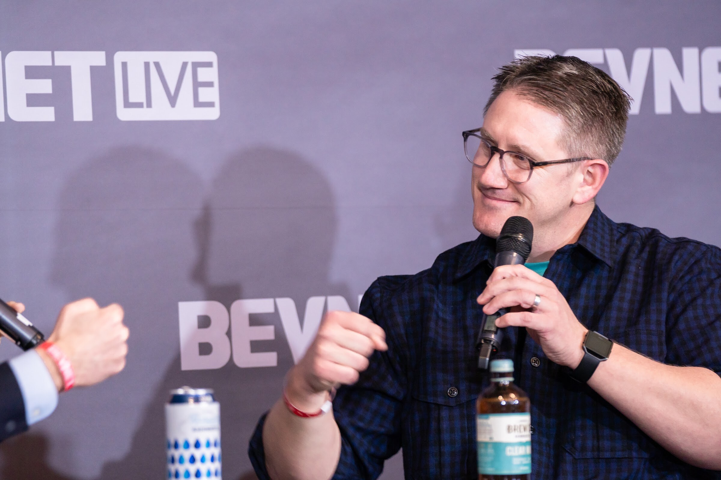 A man with glasses in a navy plaid shirt holds a microphone and smiles, with a can and a bottle on the table in front of him.