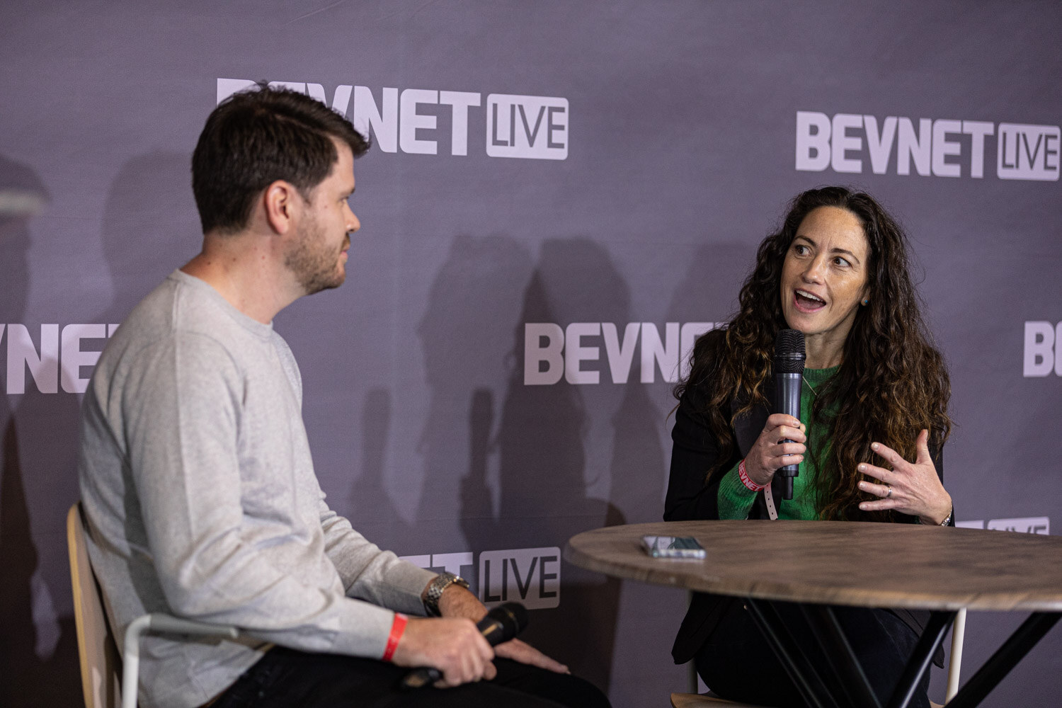 A woman in a green top with long curly hair speaks into a microphone and gestures, across a round table from a man in a gray sweater.