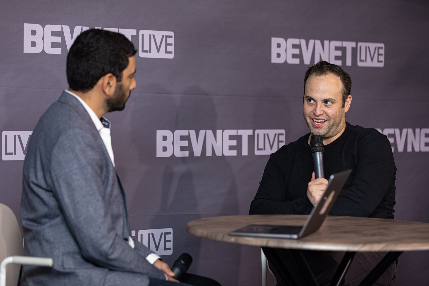 A man in a gray blazer faces a smiling man in a black sweater who holds a microphone, with a laptop on the round table between them.