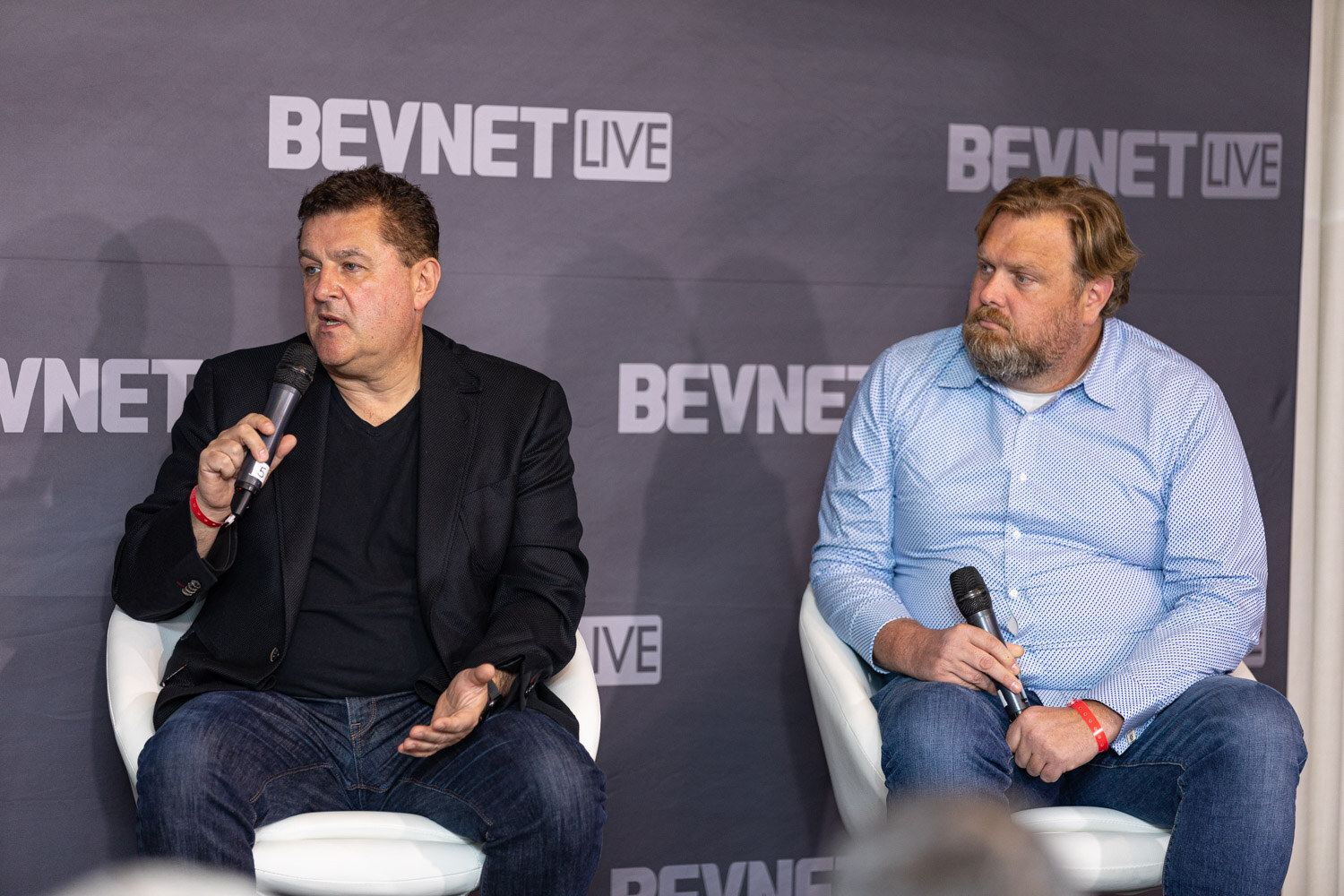 A man in a black blazer gestures while speaking next to a bearded man in a light blue checked shirt, both holding microphones.