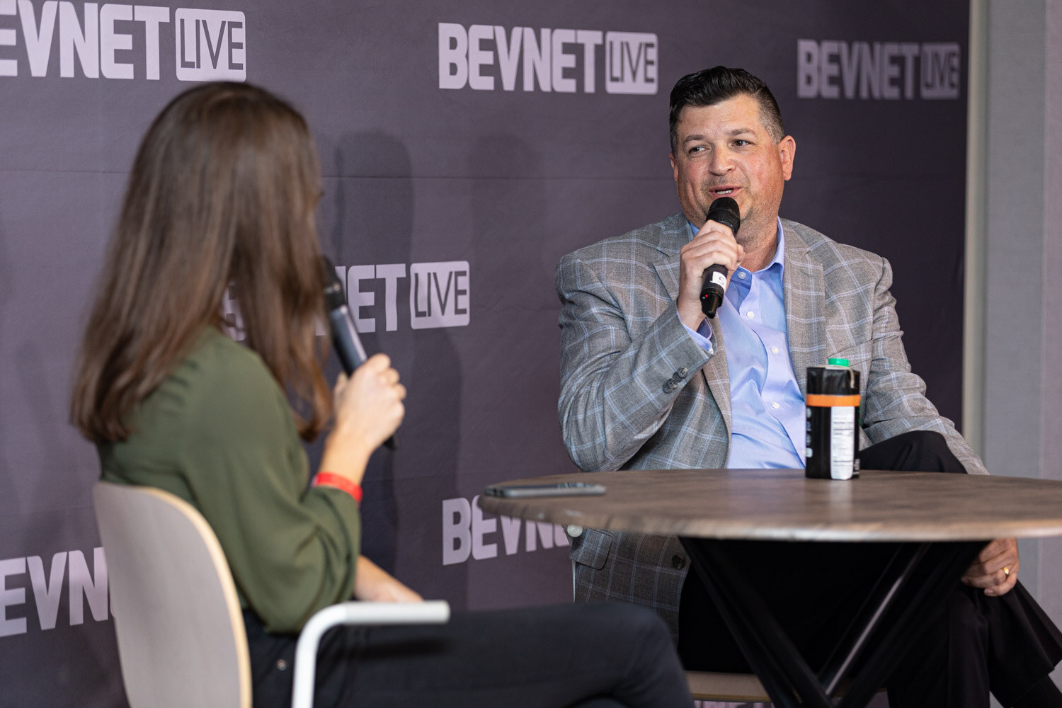 A man in a gray plaid blazer and light blue shirt holds a microphone across a round table from a woman with her back to the camera.