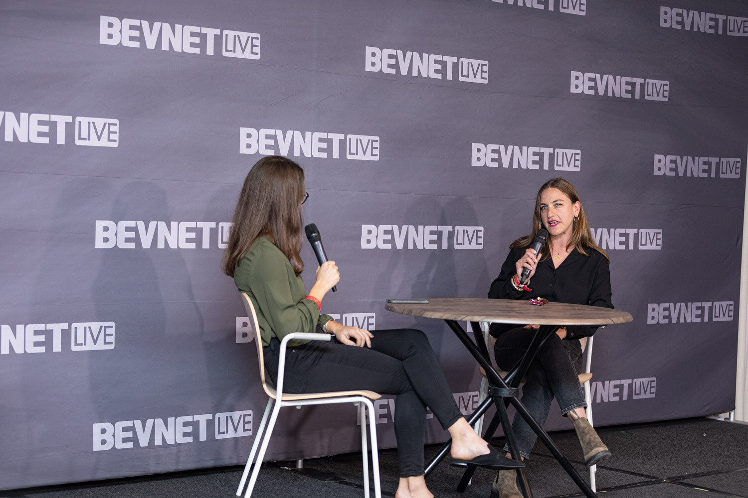 Two women seated at a small round table in front of a backdrop covered with BevNET Live logos, one holding a microphone.