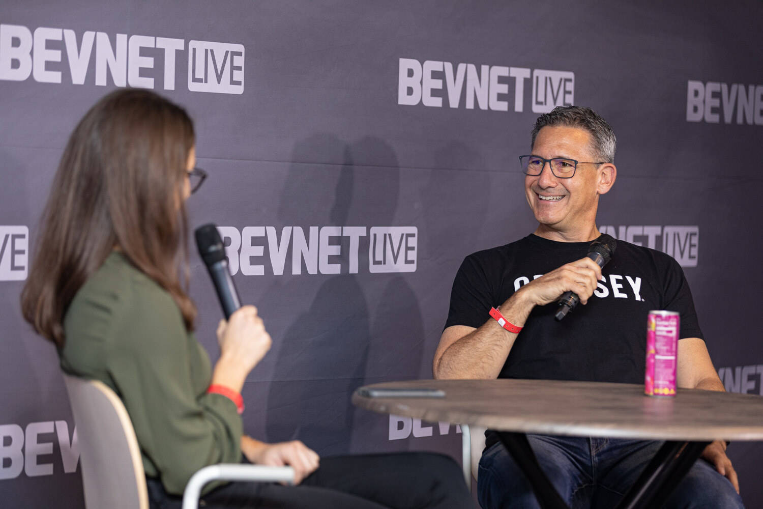 A man with glasses in a black T-shirt holds a microphone and smiles, with a pink can on the table.