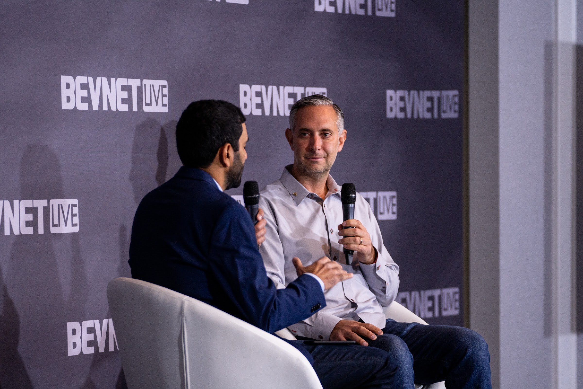 Two men sit in white chairs in front of a BevNET Live backdrop, a man in a navy blazer with his back partly to the camera and a man in a light gray shirt holding a microphone.