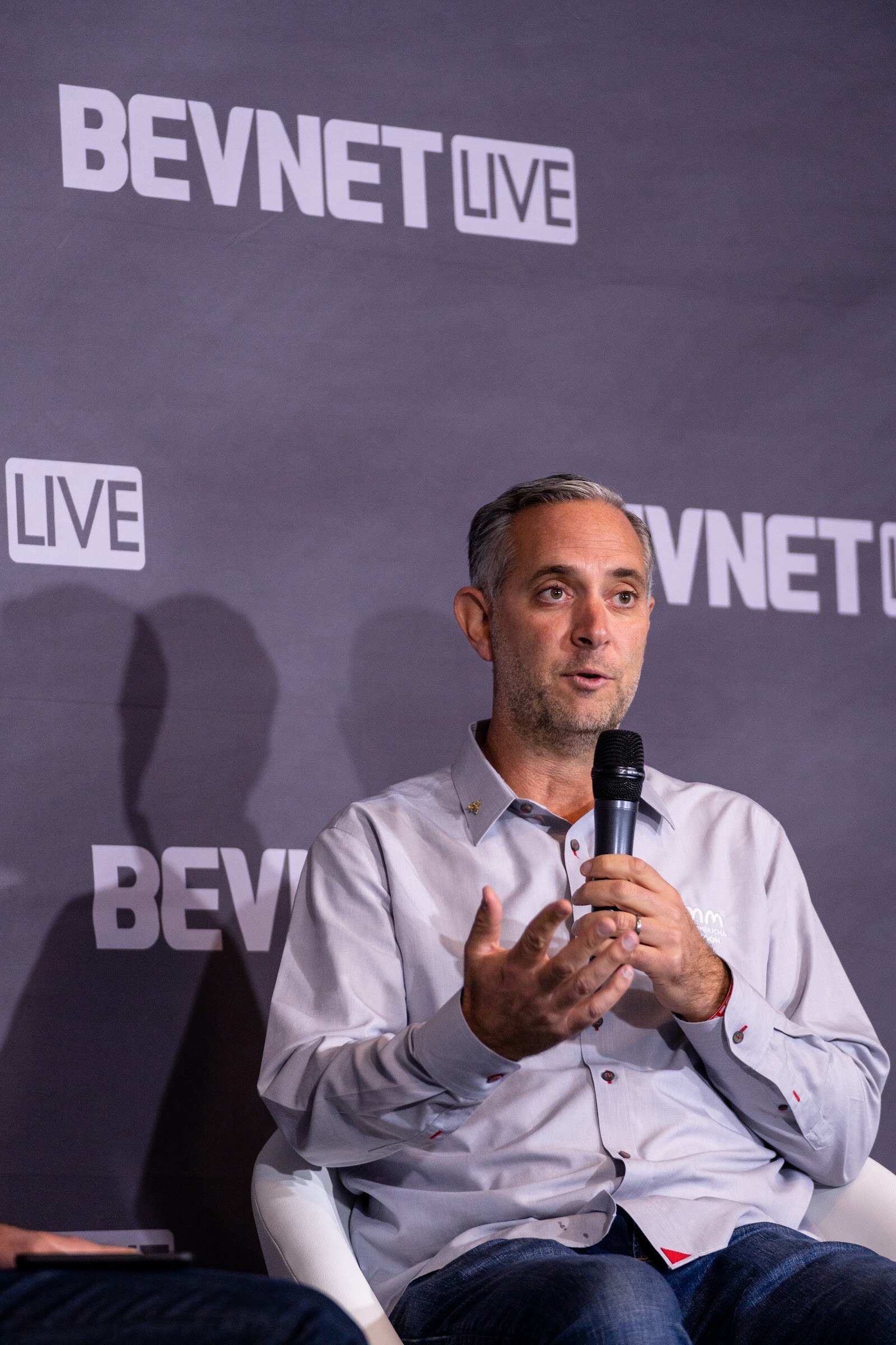 A man with gray-flecked hair in a light button-up shirt holds a microphone and gestures with open hands in front of a BevNET Live backdrop.