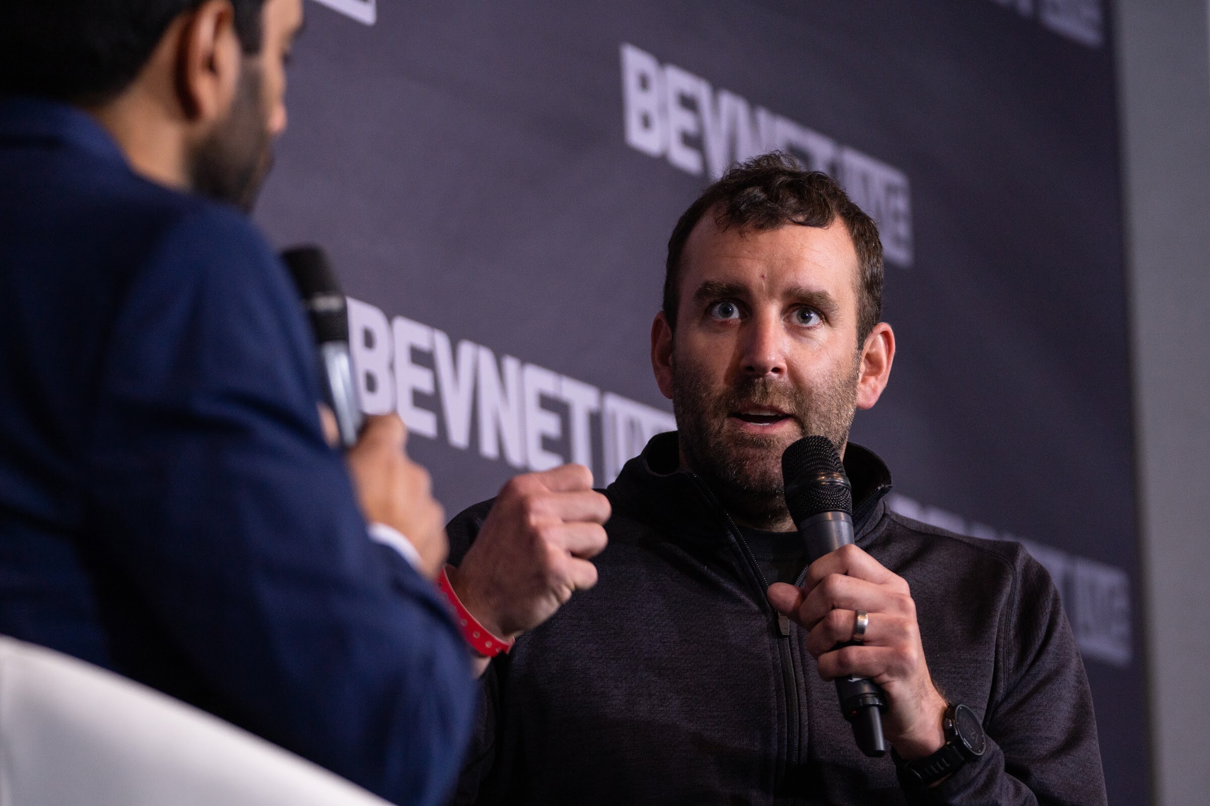 A bearded man in a dark quarter-zip holds a microphone and looks up while the interviewer's arm is blurred in the foreground.