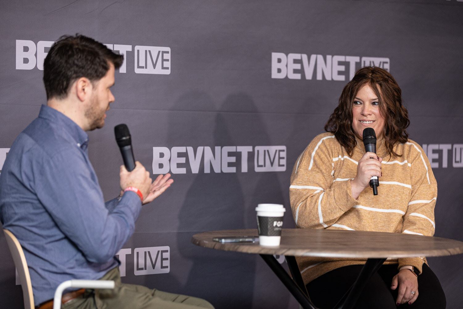 A man in a blue shirt holds a microphone toward a smiling woman in a mustard striped sweater at a round wooden table with a coffee cup.
