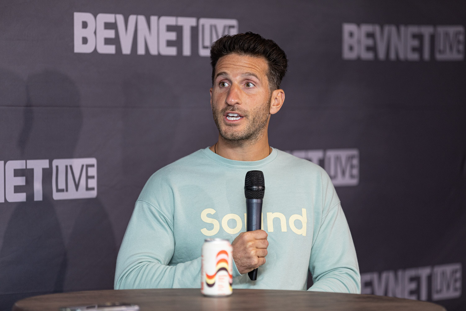 A man in a mint-green sweatshirt reading Sound holds a microphone, with a small can on the table in front of him.