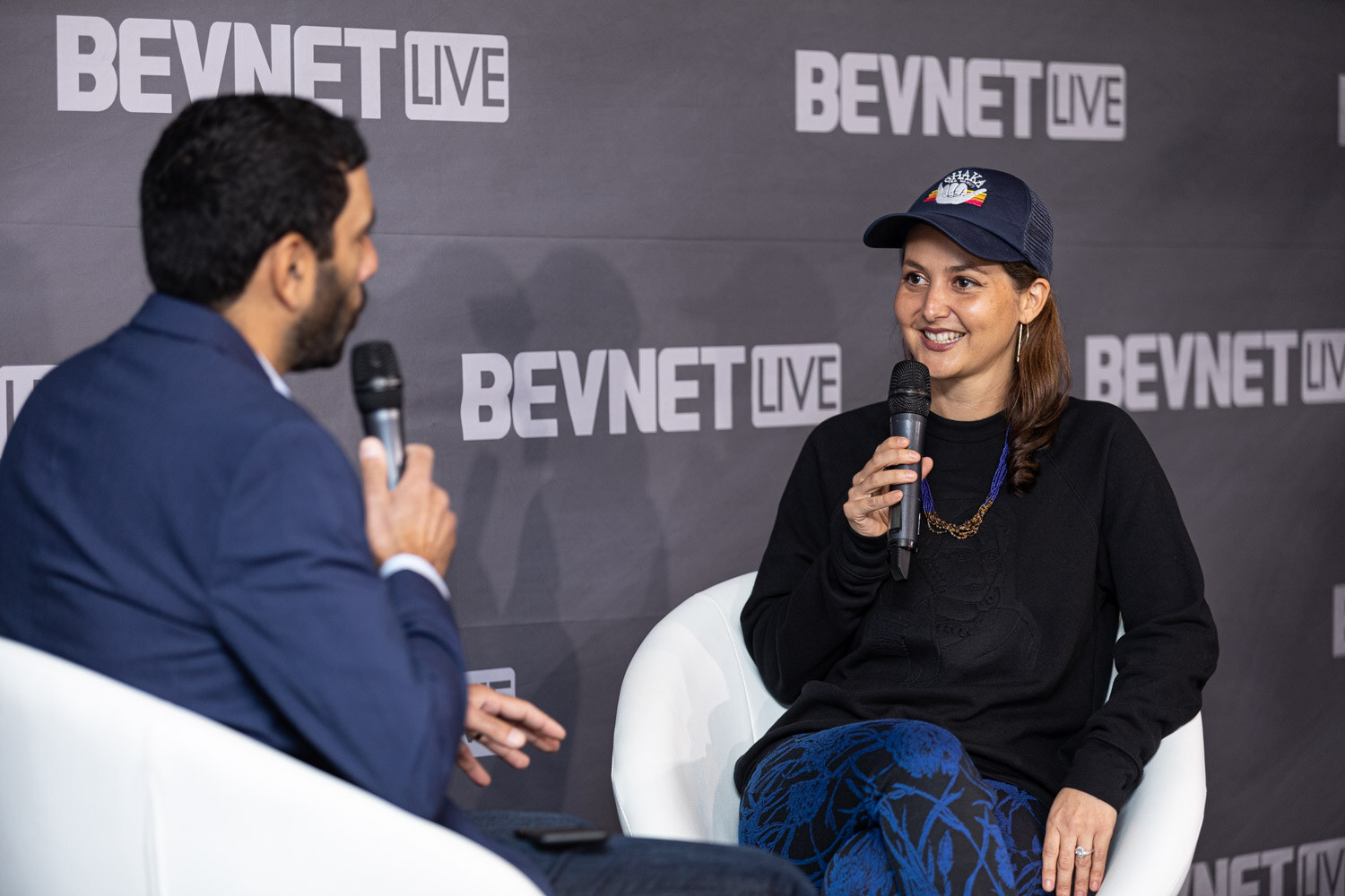 A smiling woman in a navy cap and black sweatshirt holds a microphone facing a man in a navy blazer seated in a white chair.
