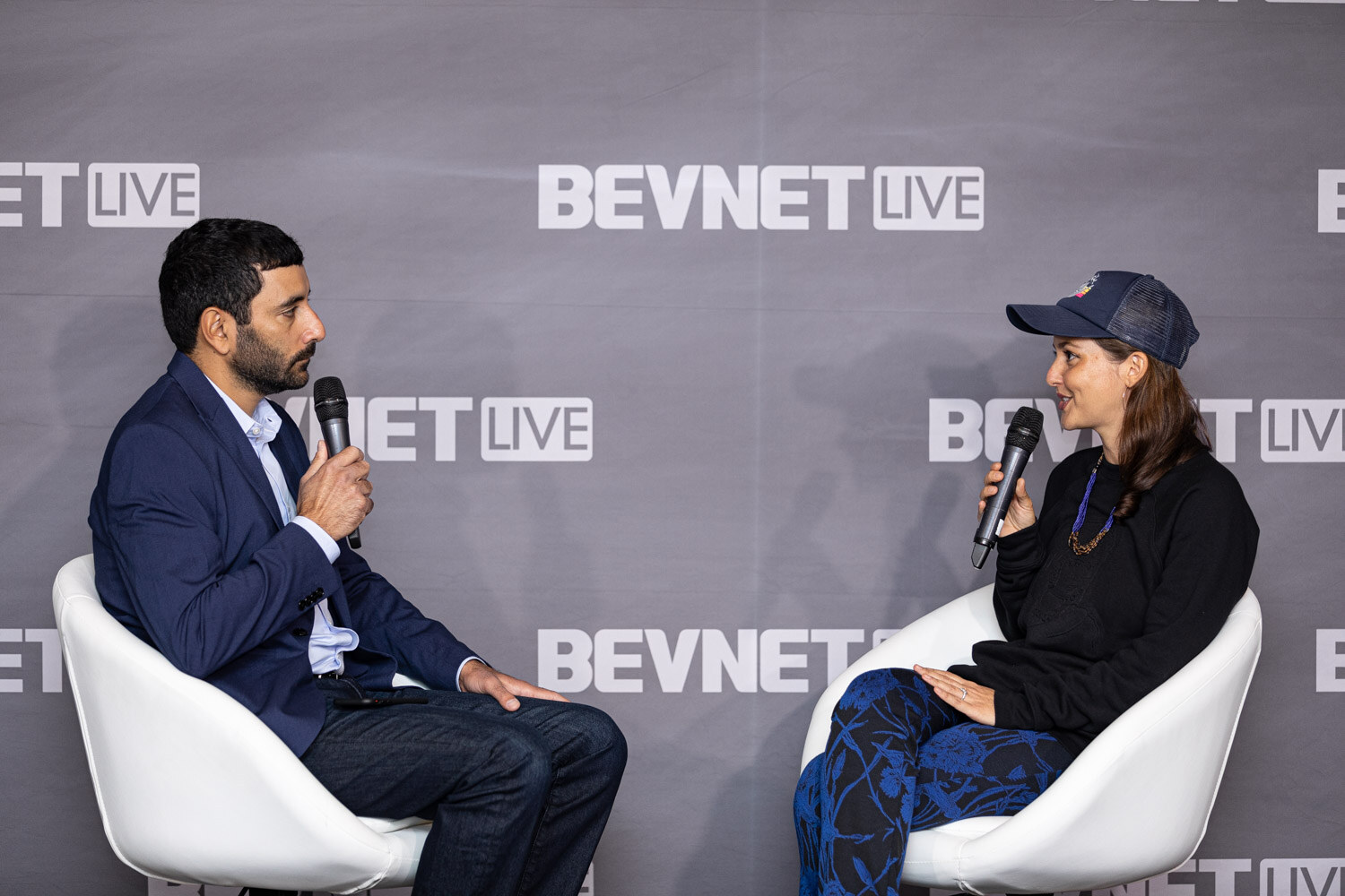 A man in a navy blazer and a woman in a navy cap and dark top sit in white chairs holding microphones in front of a BevNET Live backdrop.