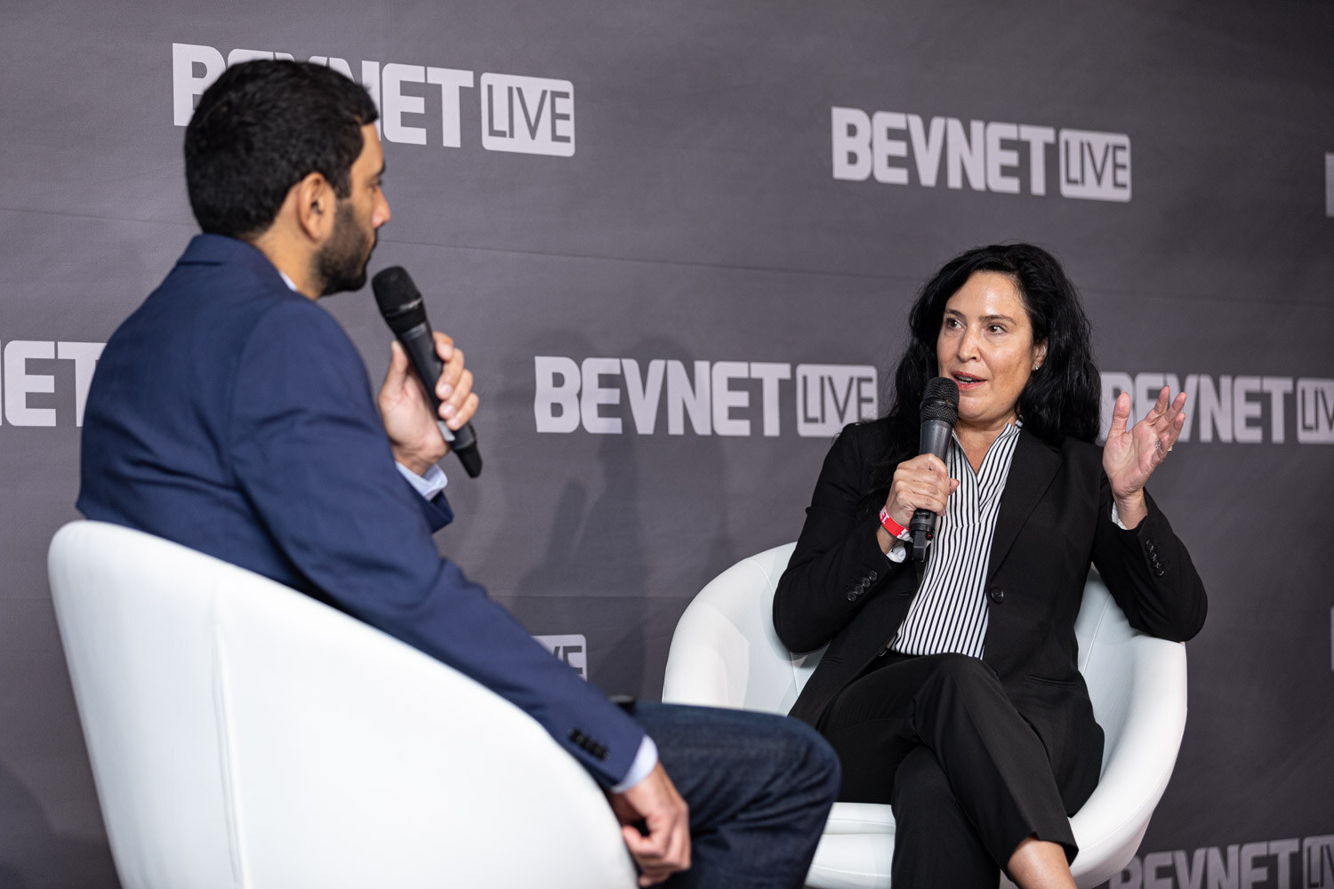 A woman with long dark hair in a black blazer and striped shirt raises one hand as she speaks, seated opposite a man in a navy blazer.