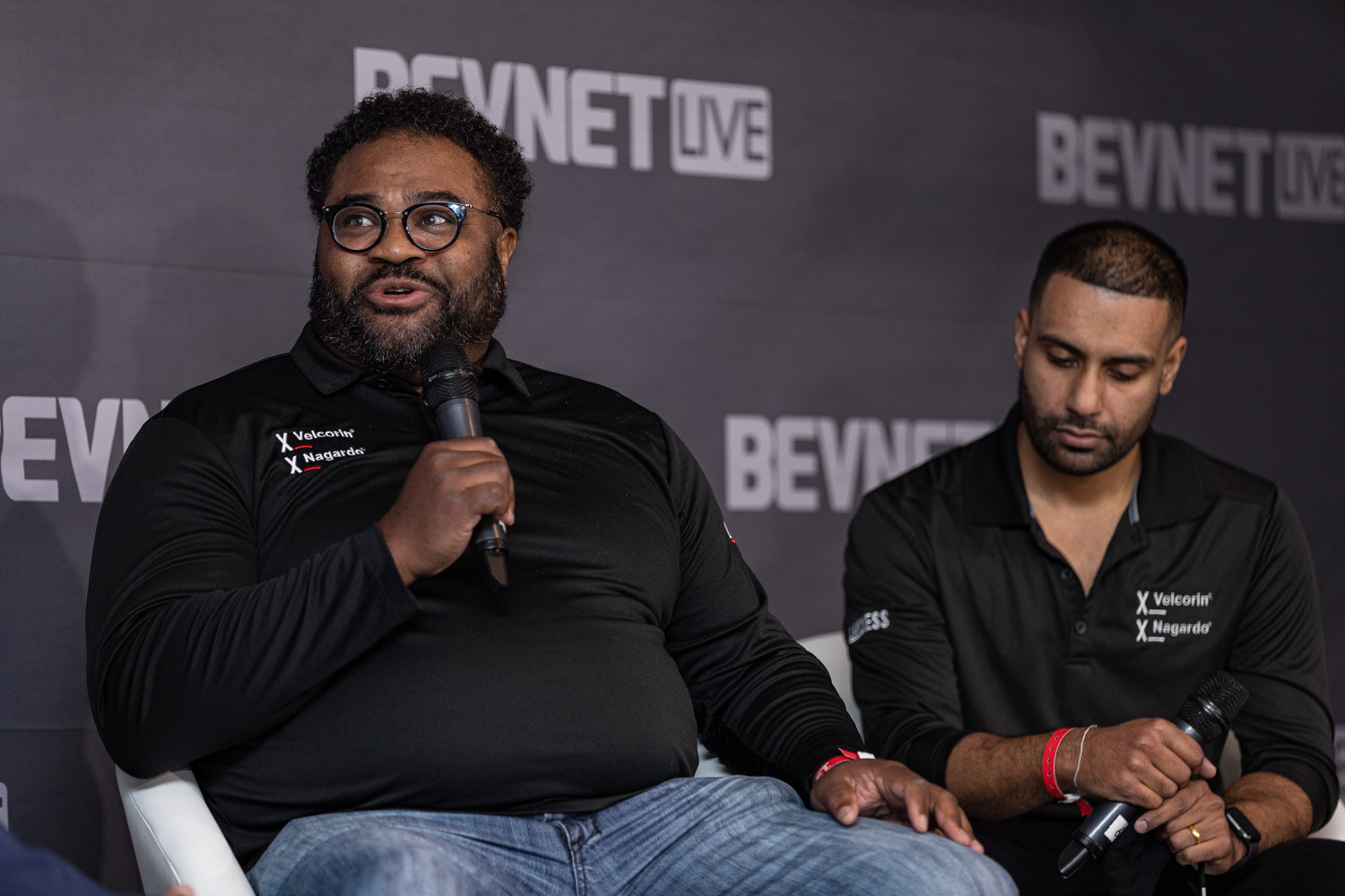 A man with glasses and a beard speaks into a microphone next to a man in a matching black polo, both seated in front of a BevNET Live backdrop.