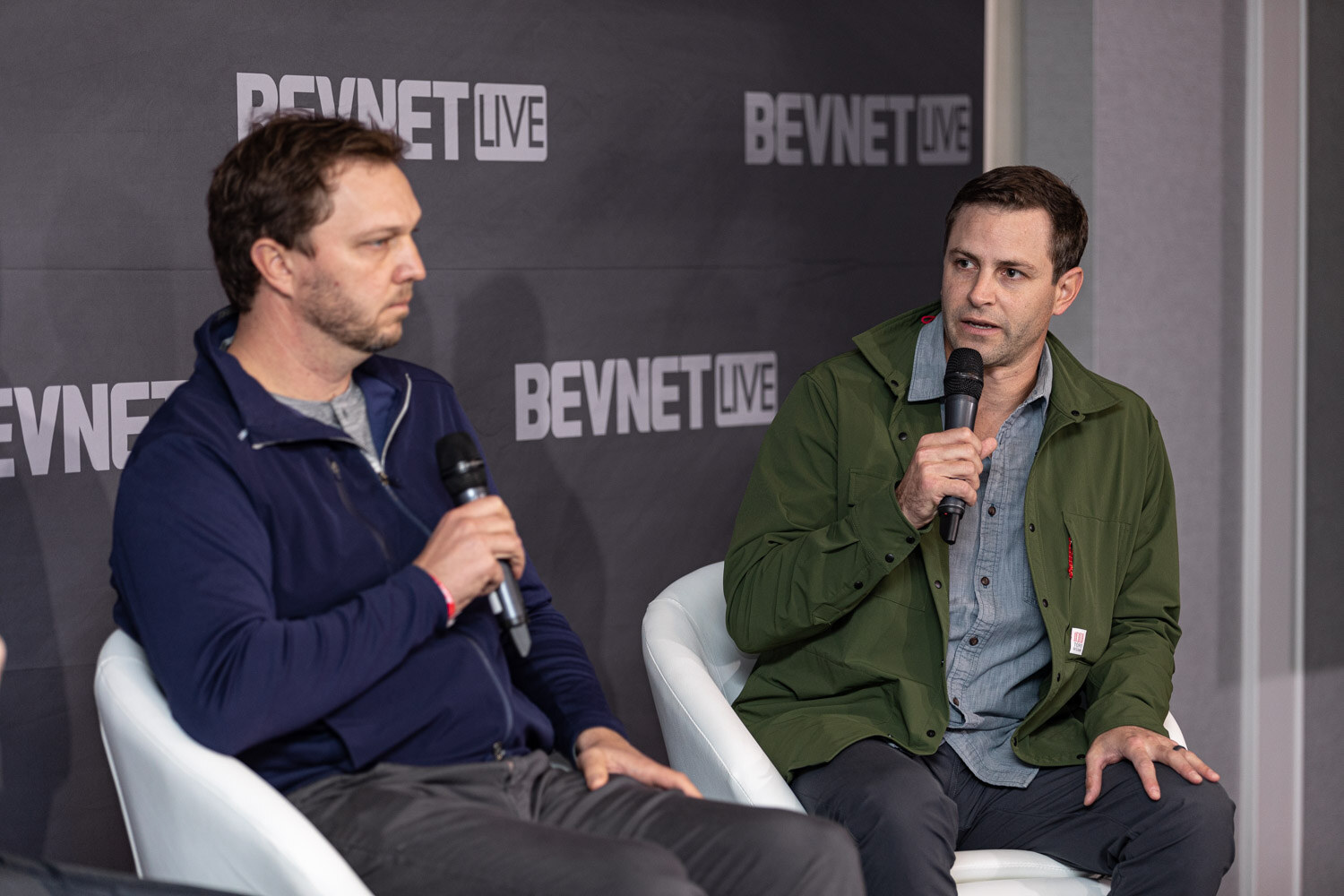 Two men sit in white chairs in front of a BevNET Live backdrop, one in a navy zip jacket and one in a green jacket, both holding microphones.