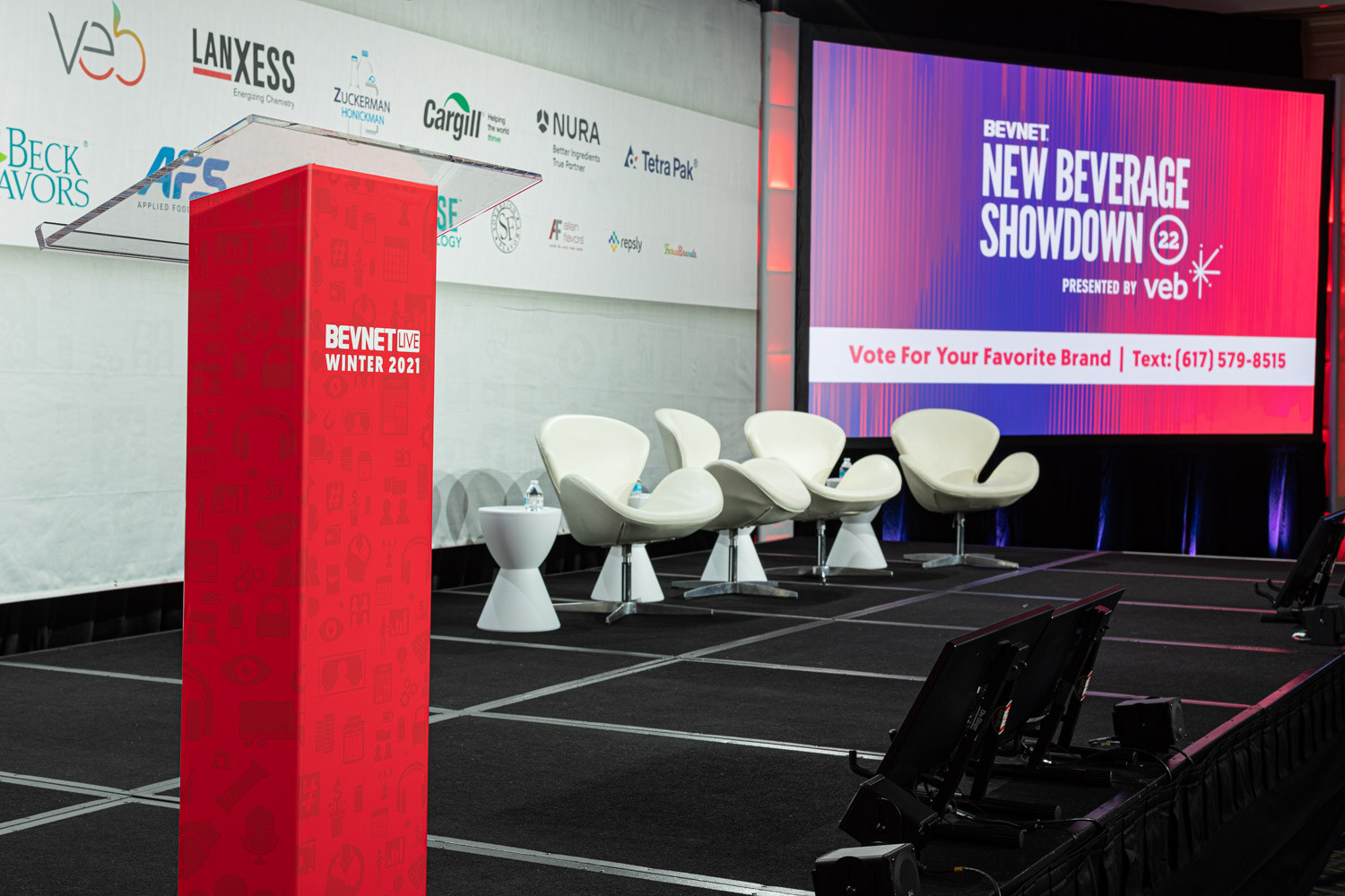 A stage with four white swan chairs, a red BevNET Live Winter 2021 podium and a large screen reading New Beverage Showdown 22 with a vote text number.