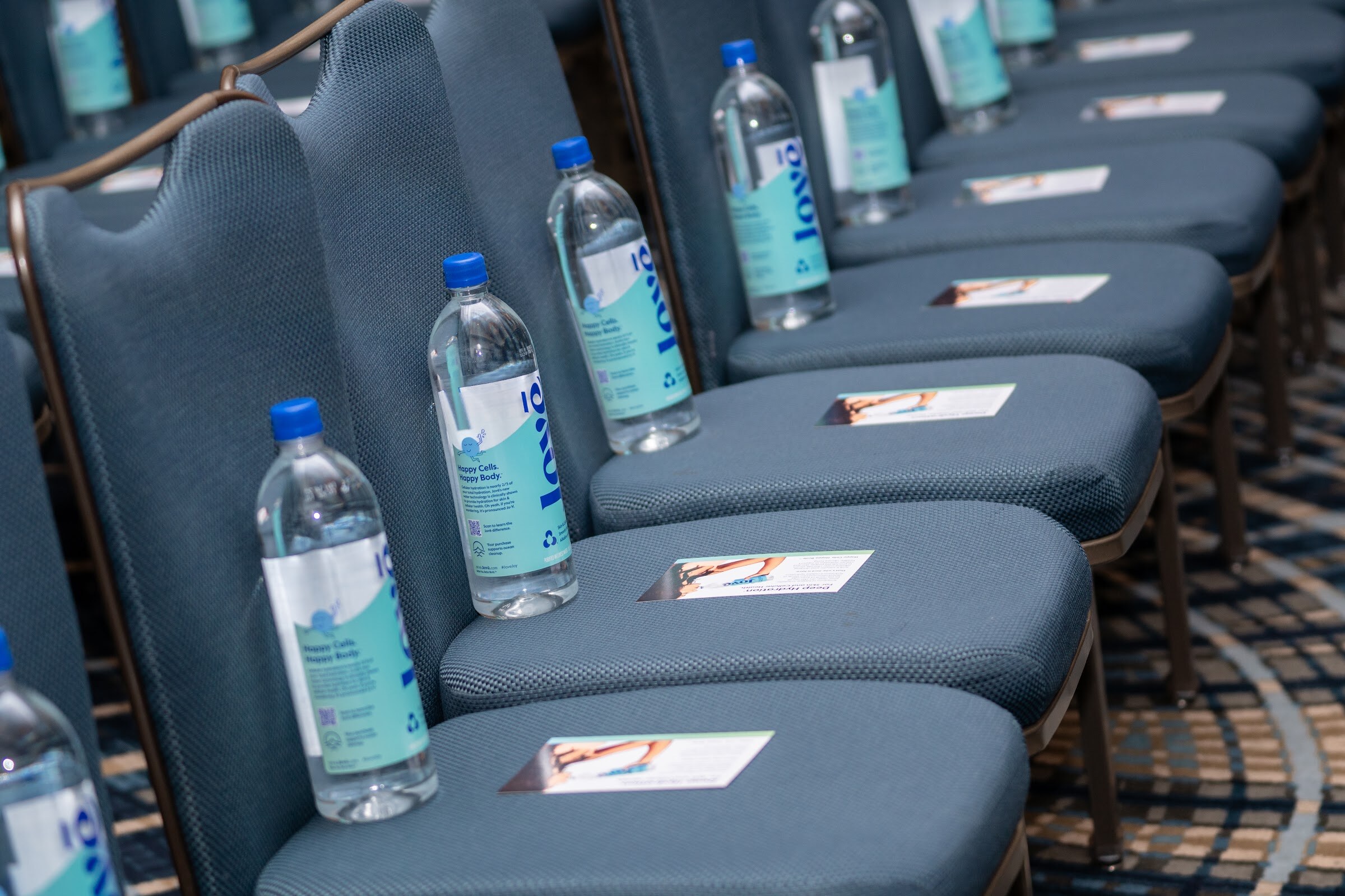 A row of teal chairs with a bottle of Jove water and a small card on each seat.