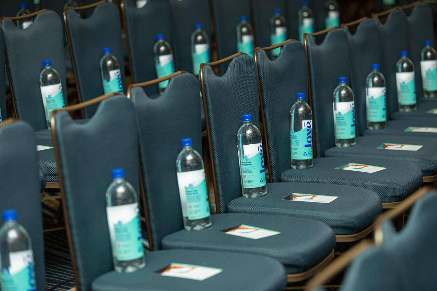 Rows of blue chairs each with a Jove water bottle and a small card on the seat.