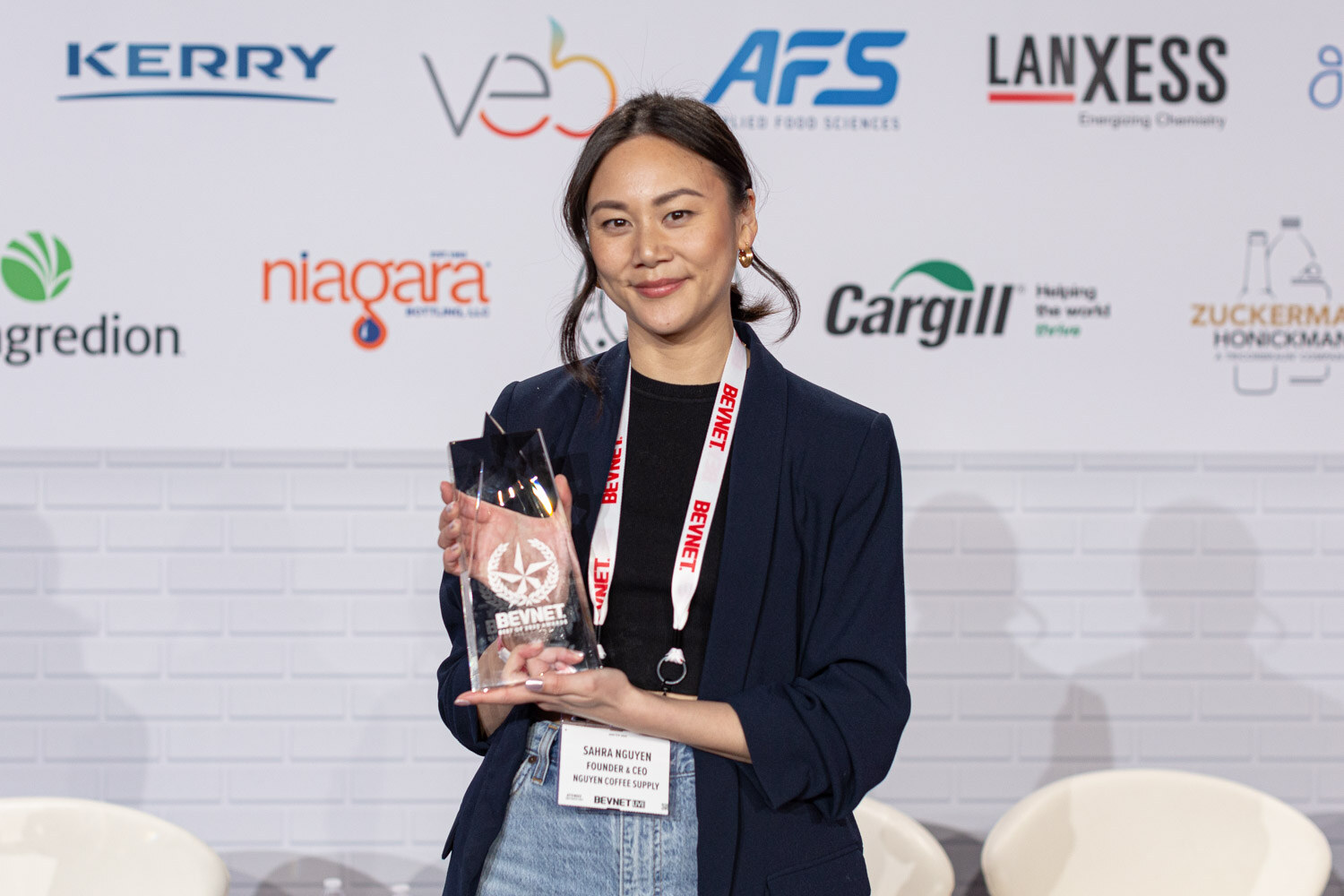 A woman in a navy blazer and black top with a BevNET lanyard smiles and holds a clear star-topped trophy in front of a white brick backdrop with sponsor logos.