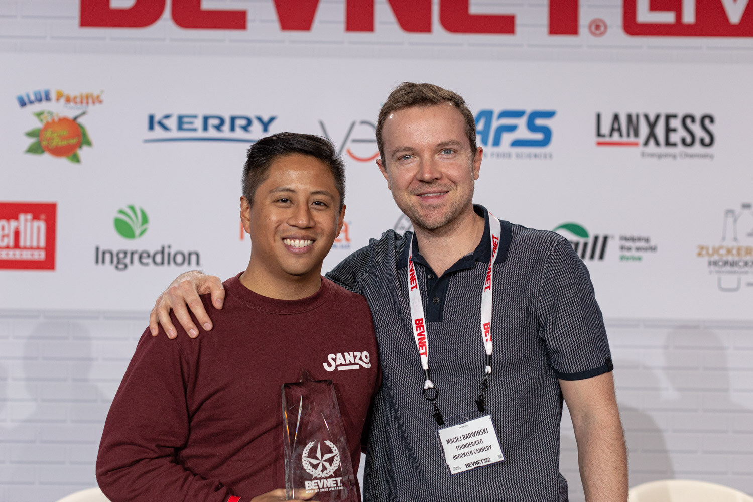 A man in a maroon Sanzo sweatshirt with his arm around a man in a navy polo, who holds a clear trophy, in front of a sponsor wall.