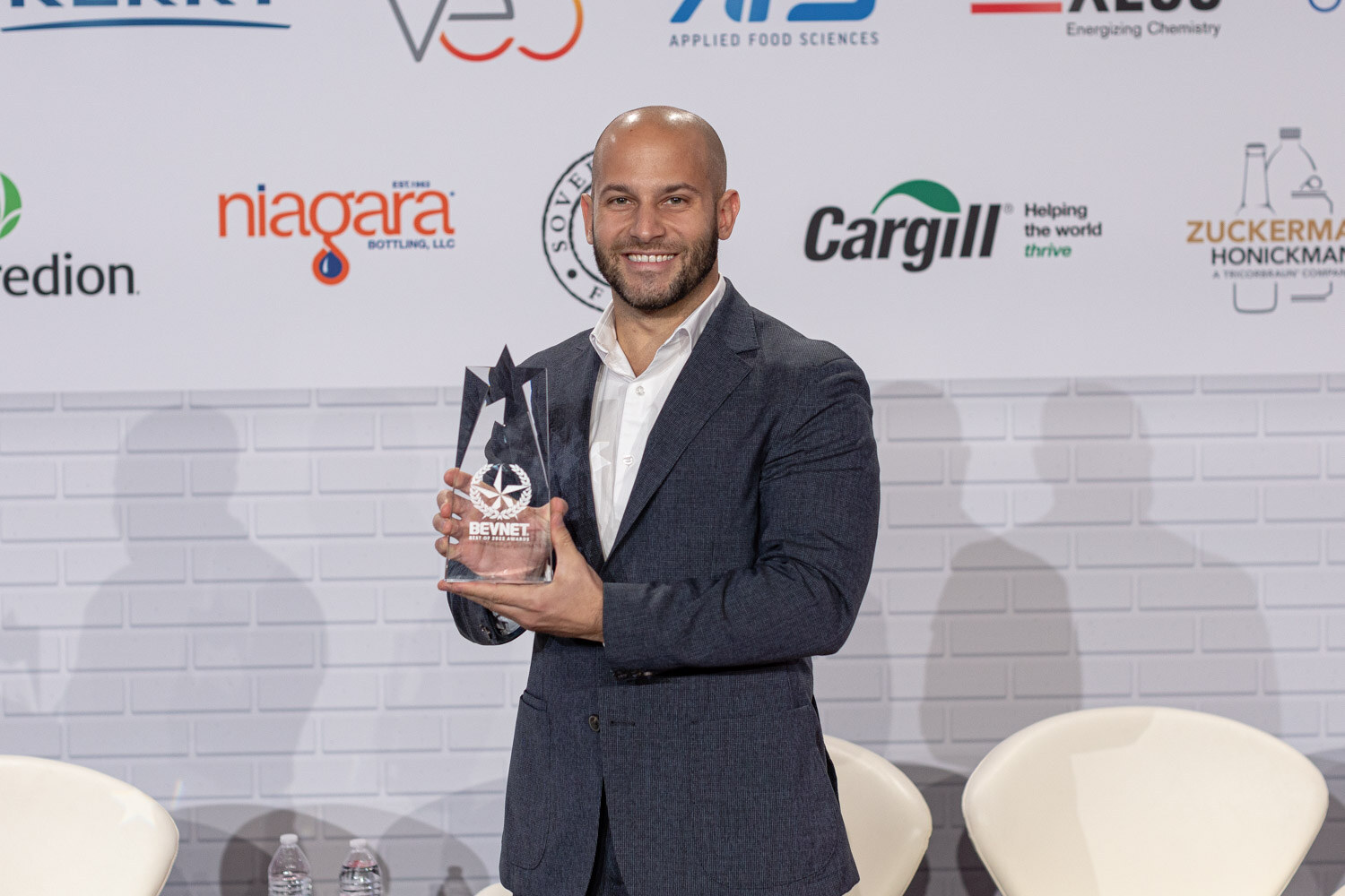 A man in a navy blazer and white shirt holds a clear star-shaped trophy in front of a sponsor wall.