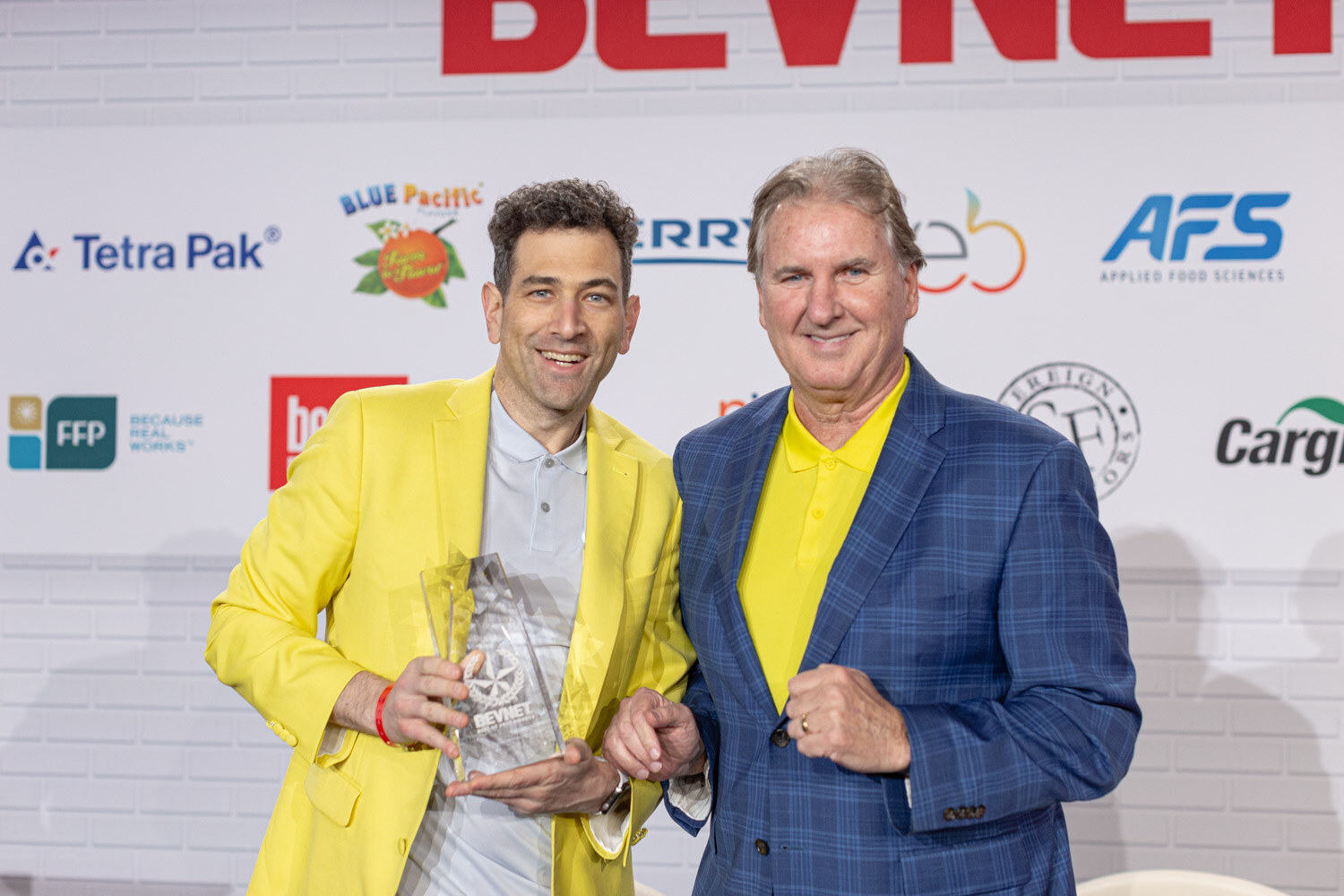 A man in a bright yellow blazer and a man in a blue plaid blazer with a yellow shirt hold a clear trophy together in front of a sponsor wall.