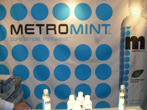 A MetroMint backdrop with blue dots and the tagline "pure. simple. mintwater." above a counter holding several bottles of MetroMint water.