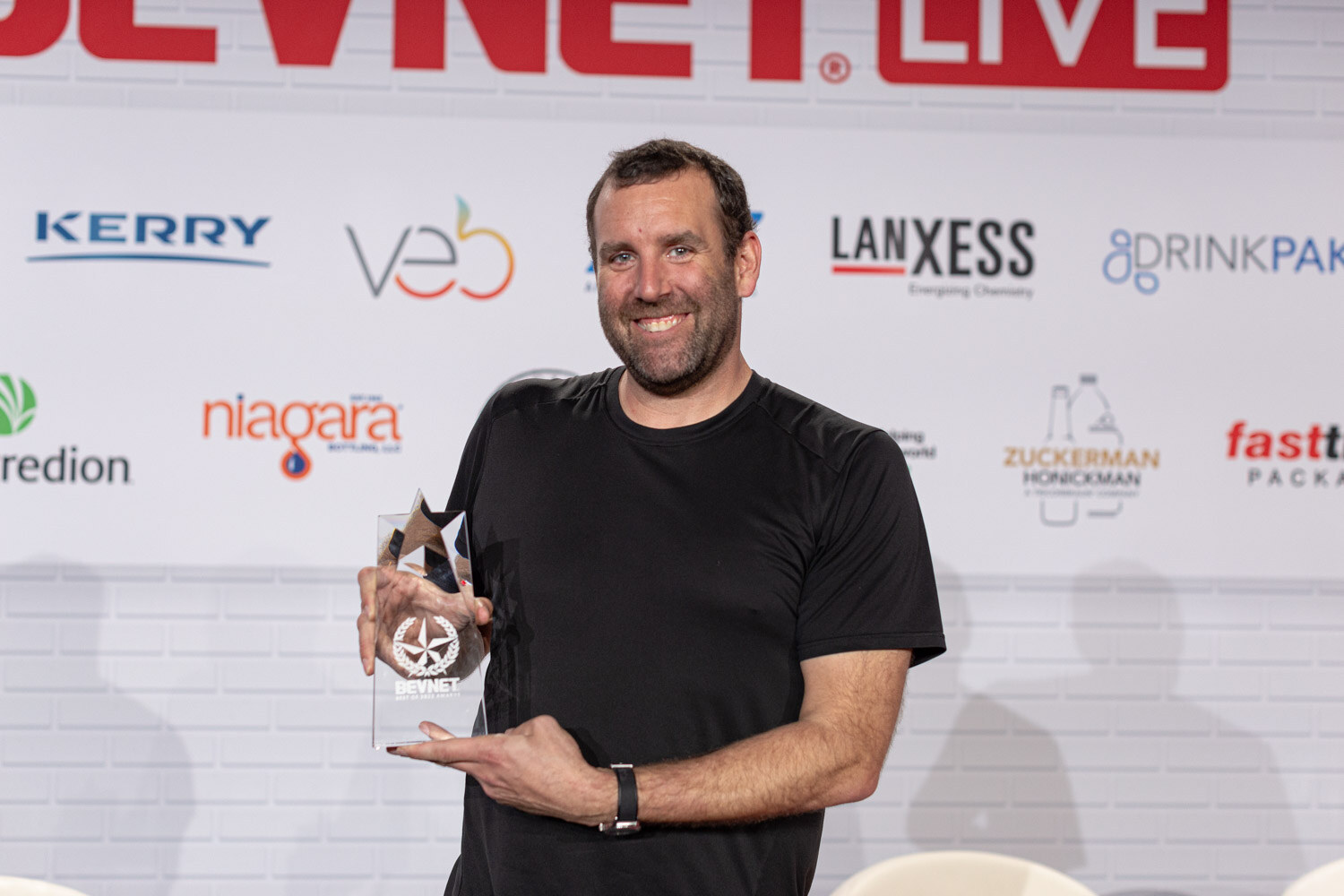 A man in a black t-shirt holds a clear trophy in front of a white brick wall with sponsor logos.