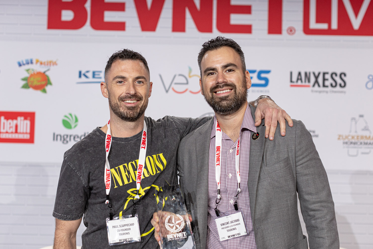 Two smiling men with BevNET lanyards, one in a Nirvana-style t-shirt and one in a gray blazer, pose with a clear trophy in front of a sponsor wall.