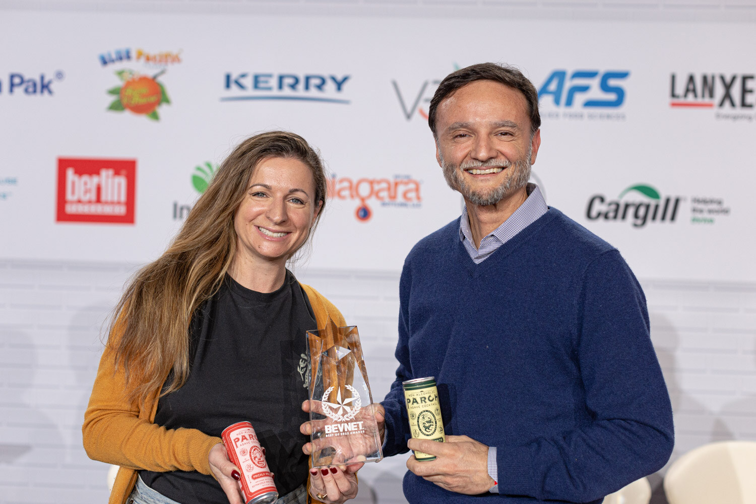 A woman in an orange cardigan holding a red can and a trophy stands beside a man in a navy sweater holding a green can.