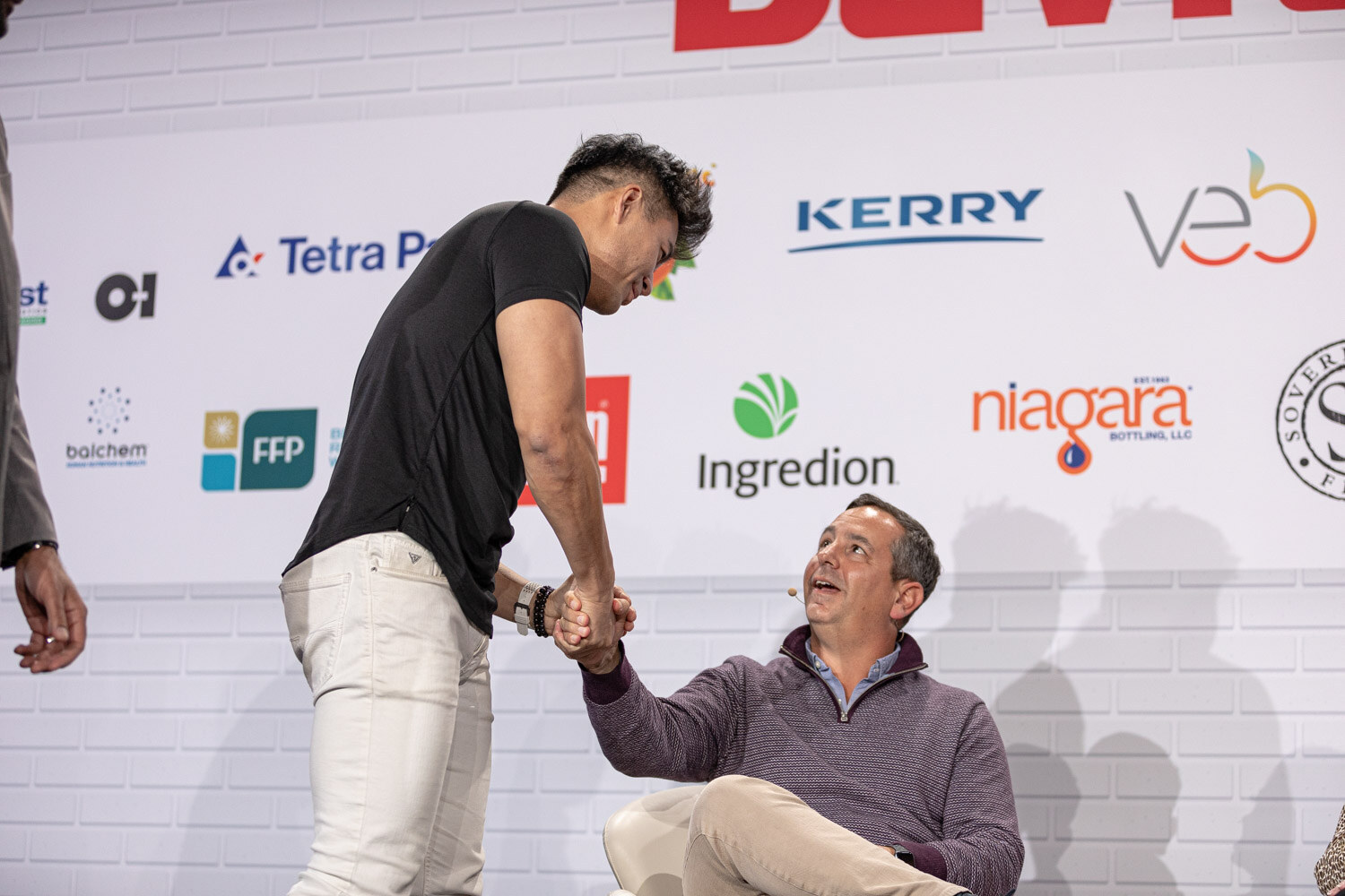 A man in a black t-shirt and white pants leans down to shake hands with a seated man in a purple sweater on stage.