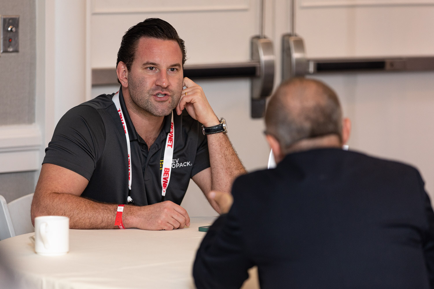 A man in a dark polo with a BevNET lanyard leans on a table and talks to another person seen from behind in a hotel meeting room.