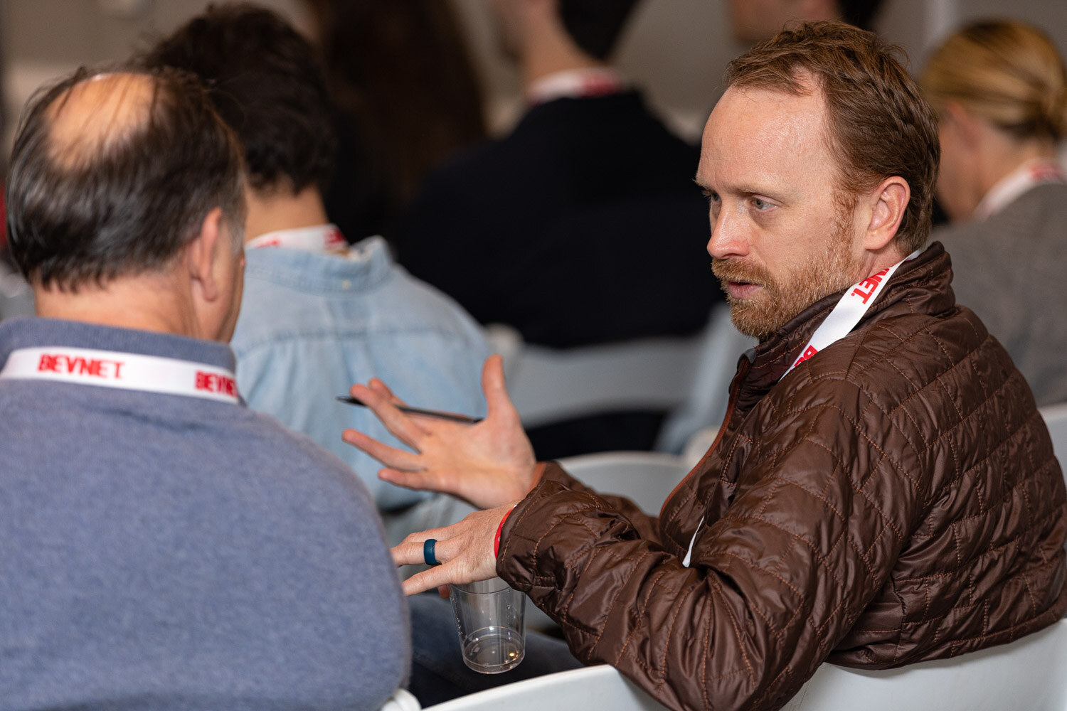 A man in a brown puffer jacket and BevNET lanyard gestures as he talks with a man in a blue sweater seen from behind.
