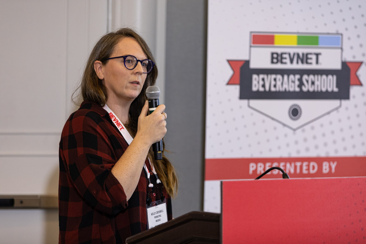 A woman in glasses and a red plaid shirt holds a microphone beside a lectern and a banner reading BevNET Beverage School Presented by.