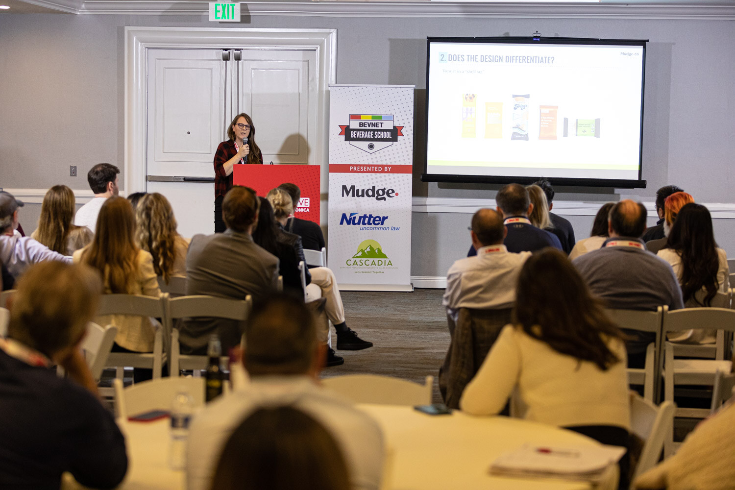 A hotel meeting room with seated attendees at tables facing a speaker at a red podium, a projection screen and a BevNET Beverage School banner.