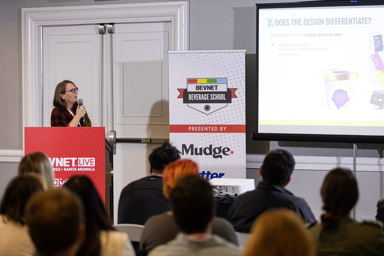 A woman at a red BevNET Live podium beside a Beverage School banner and a screen reading Does the design differentiate, with attendees in the foreground.