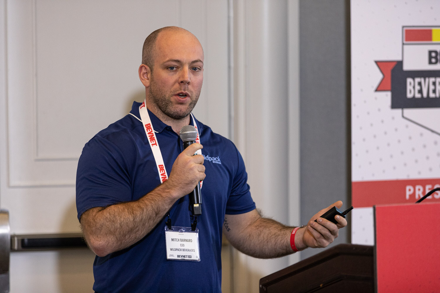 A man in a navy polo with a BevNET lanyard and Wildpack Beverages badge holds a microphone and clicker in front of a Beverage School banner.