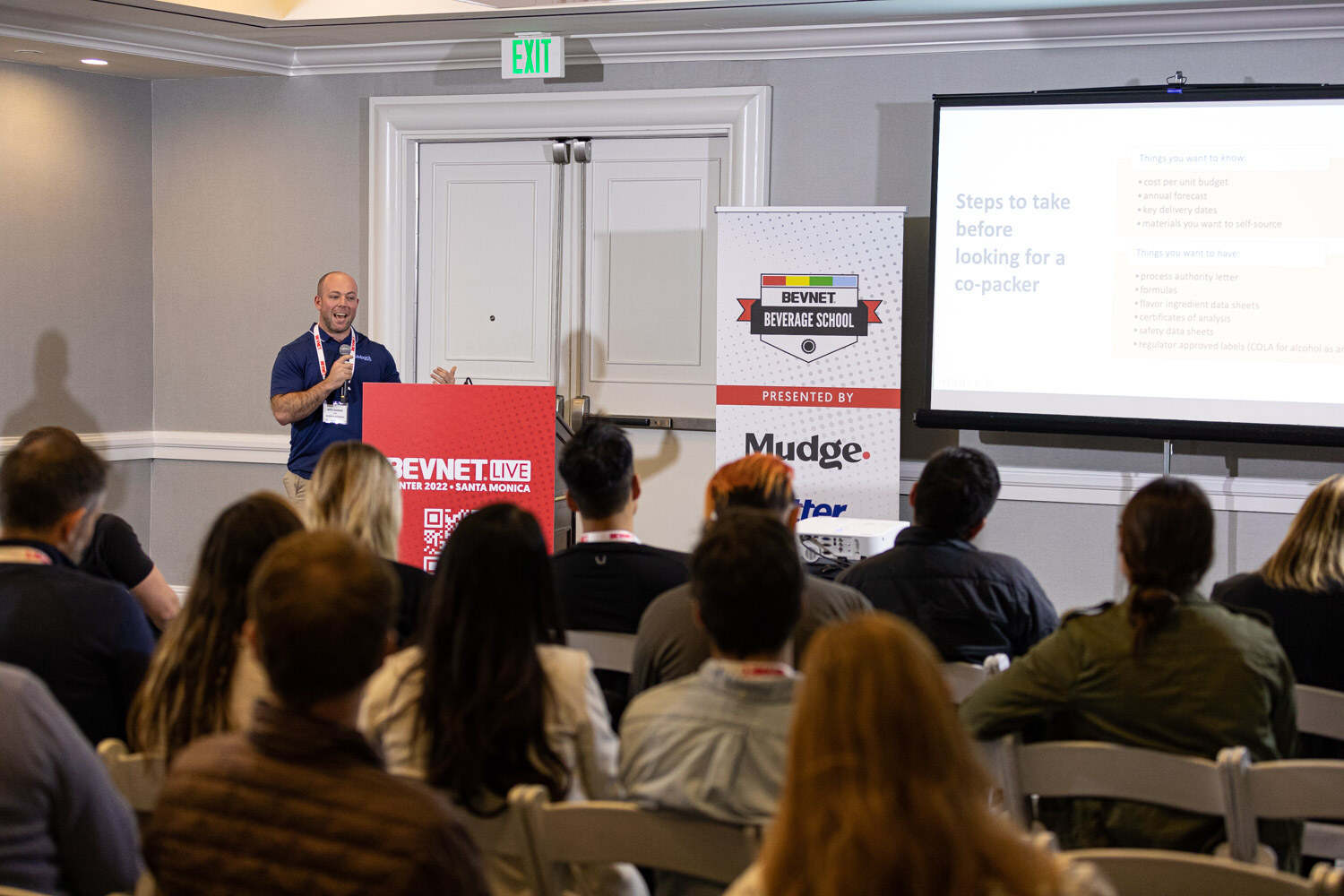 A speaker at a red podium beside a screen reading Steps to take before looking for a co-packer, with attendees seated in the foreground.