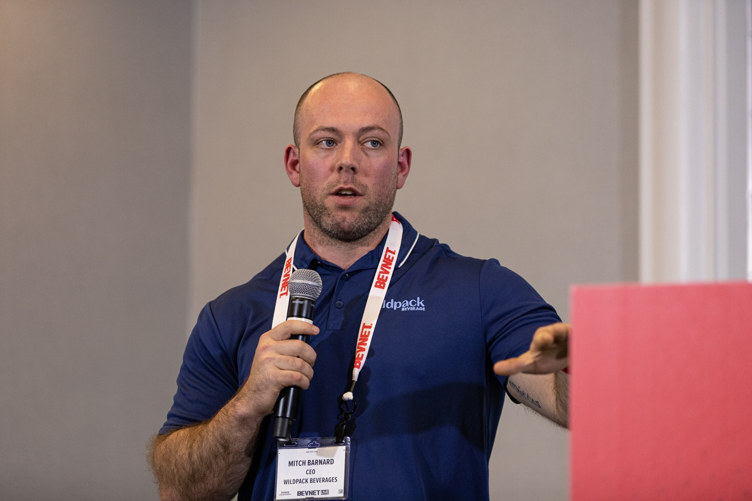 A bald man in a navy polo with a Wildpack Beverages badge and BevNET lanyard holds a microphone and points to his left.