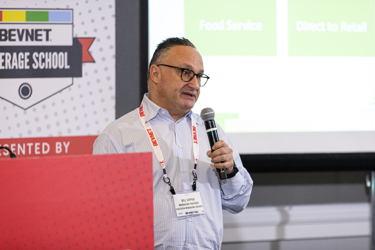 A man in a light checked shirt with a BevNET lanyard and badge holds a microphone beside a red lectern in front of a Beverage School banner.