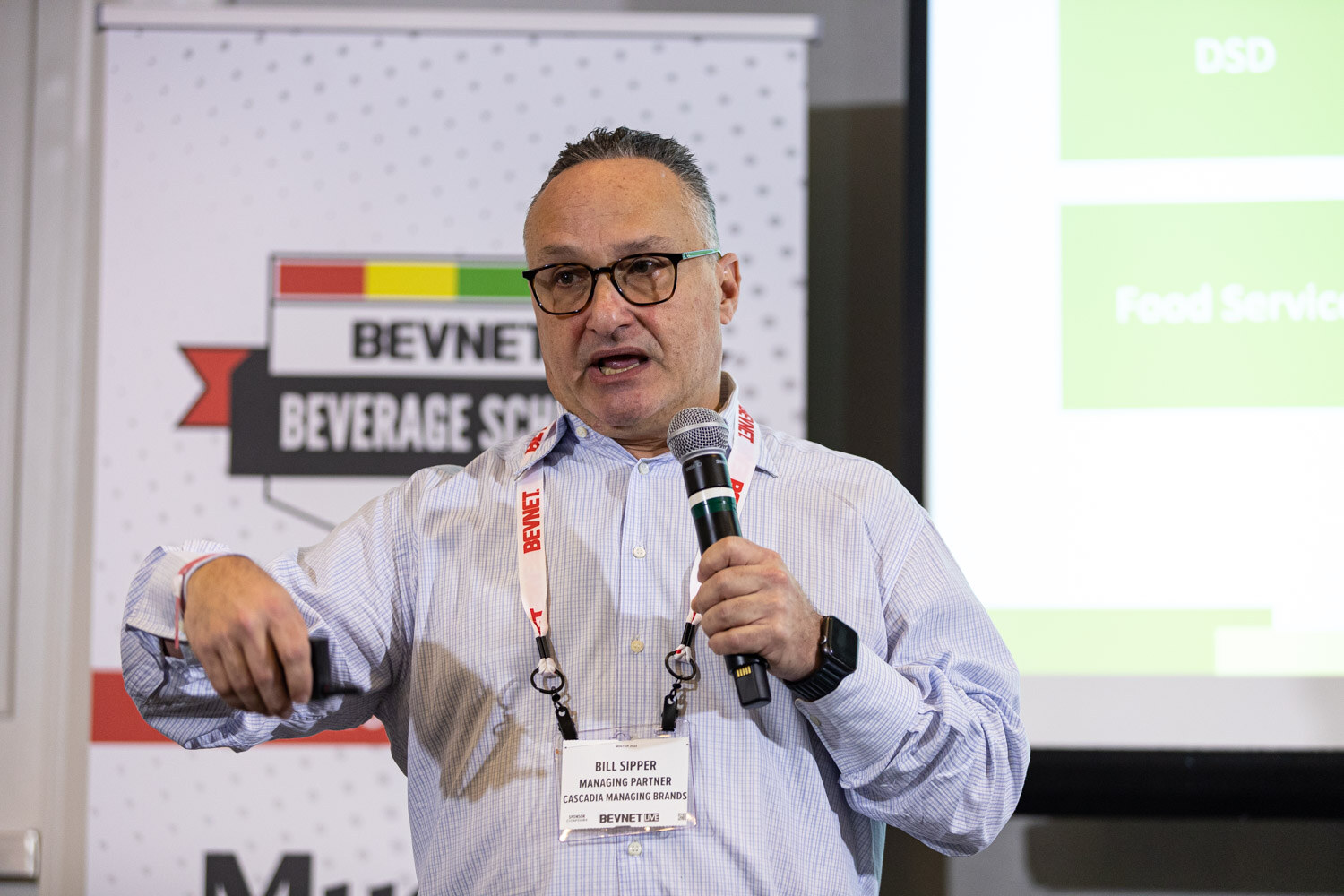 A man in glasses and a light checked shirt holds a microphone and gestures, with a slide listing Food Service and DSD behind him.