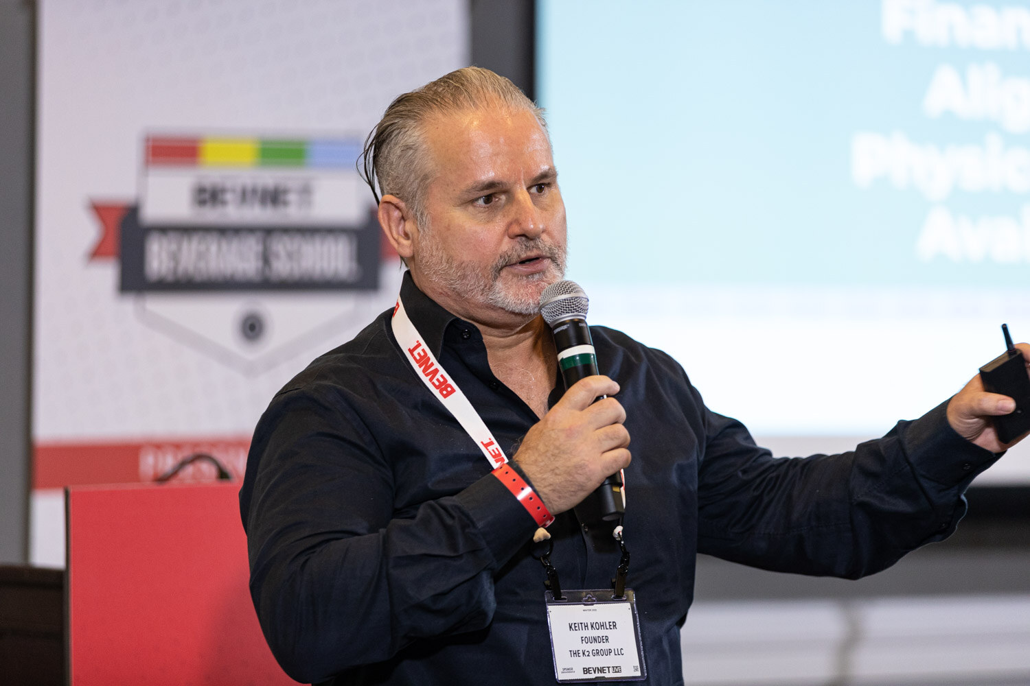 A man in a black shirt with a BevNET lanyard and badge holds a microphone and points to a screen showing a funding stages slide.