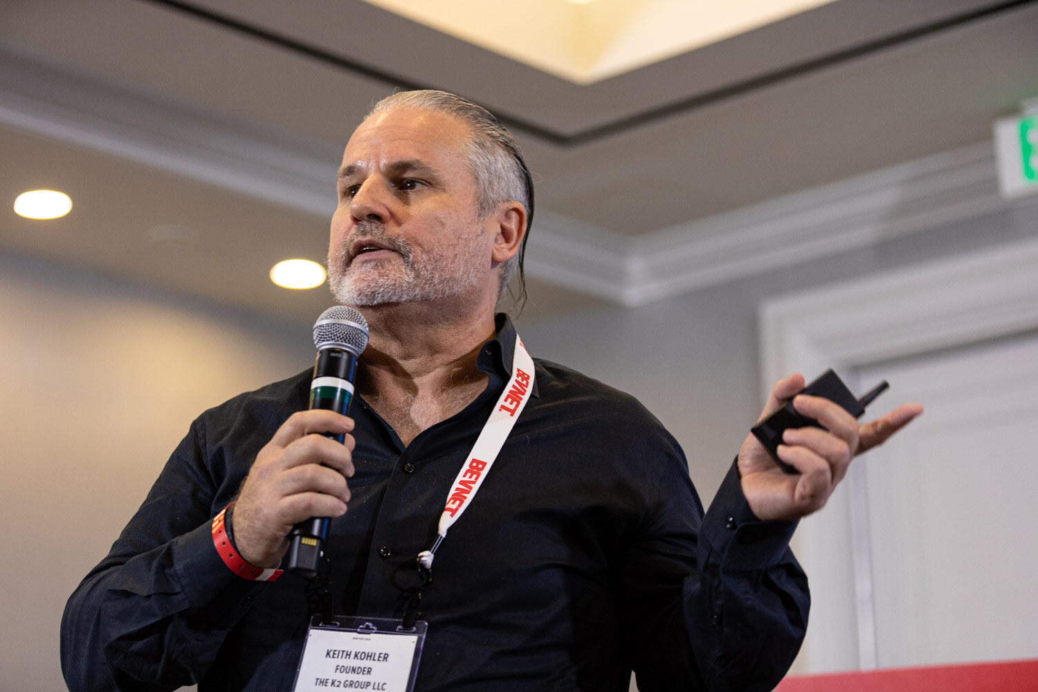 A man in a black shirt with a red BevNET lanyard speaks into a microphone while holding a clicker in the other hand.