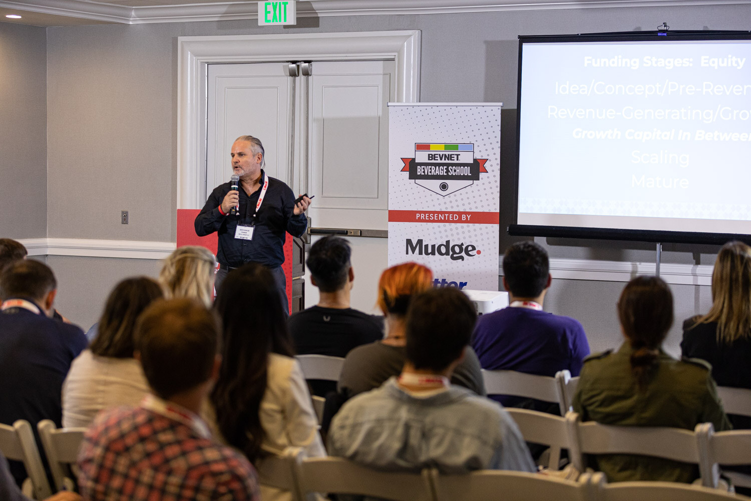 A speaker stands at the front of a meeting room beside a Beverage School banner and a screen listing equity funding stages, with attendees seated in the foreground.