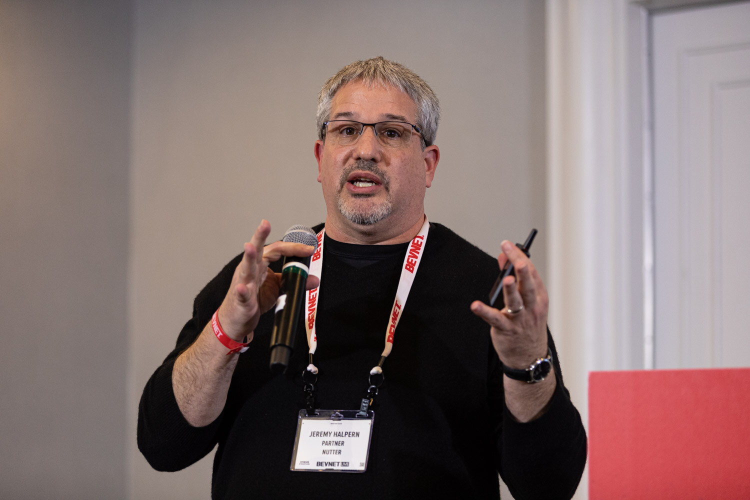 A man in glasses and a black sweater with a BevNET lanyard holds a microphone and raises both hands.