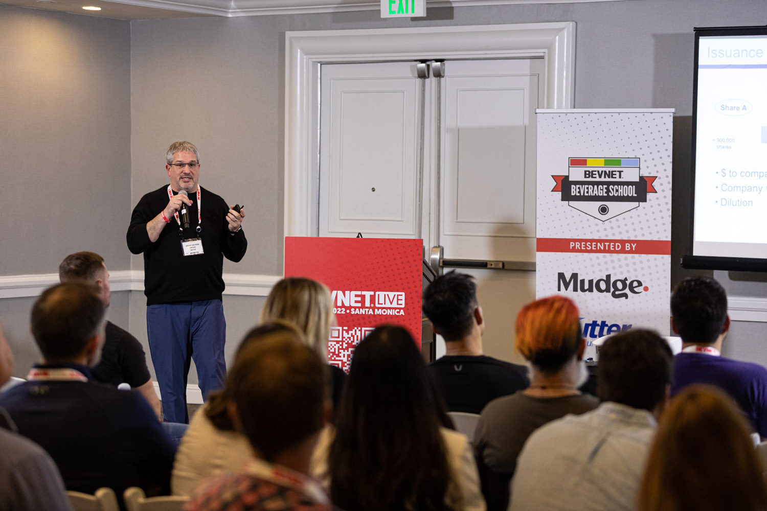 A man in a black sweater and jeans stands beside a red BevNET Live podium and Beverage School banner facing attendees seated with their backs to the camera.